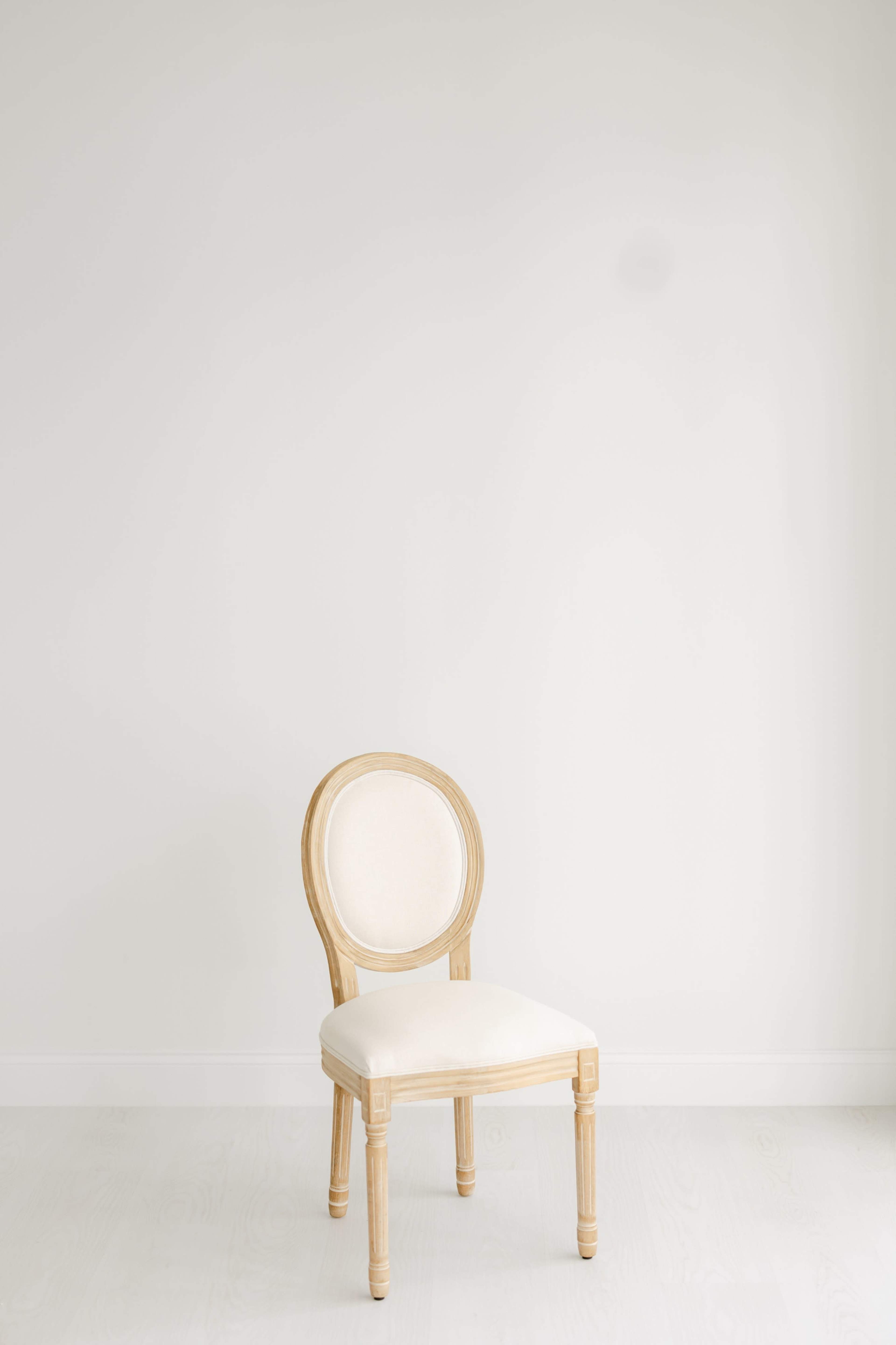 A wooden chair with a round backrest and a cream-colored seat is positioned against a blank white wall.
