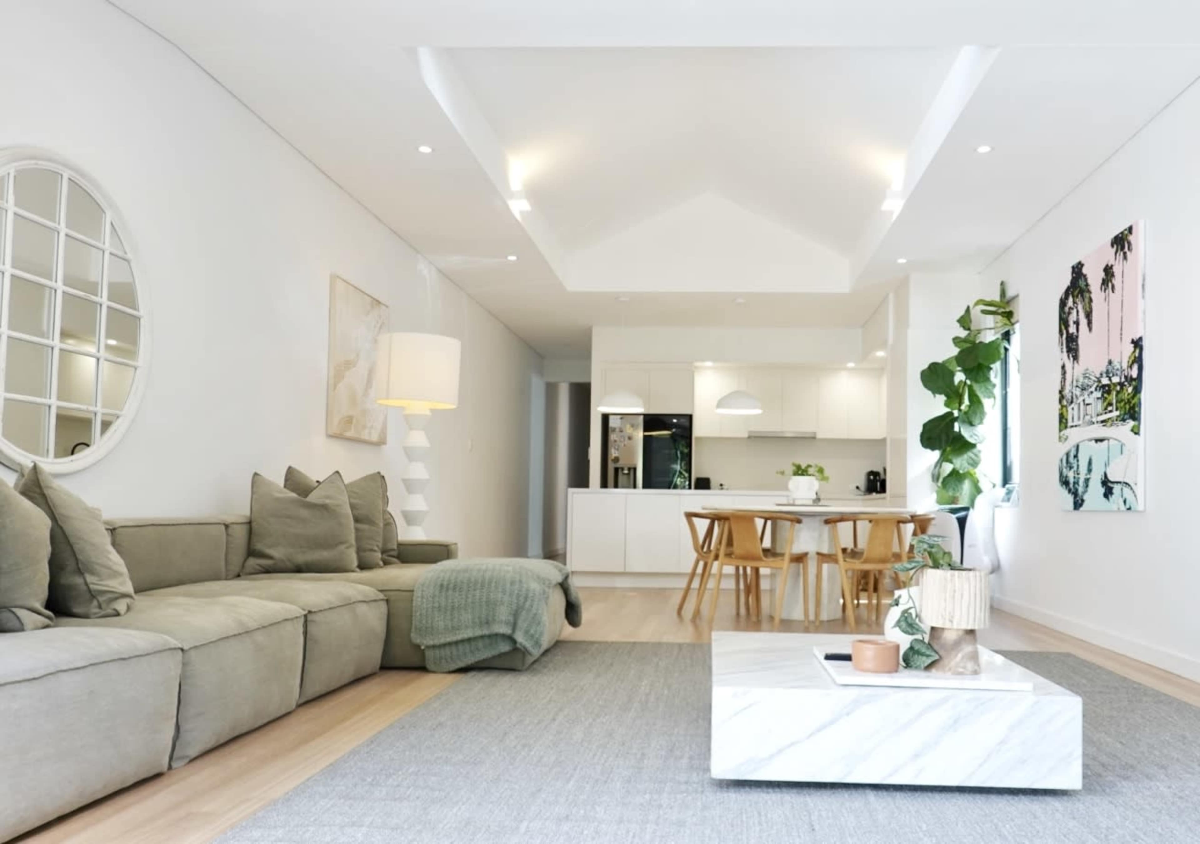 The image shows a modern living room with a sectional sofa, a marble coffee table, and a dining area featuring wooden chairs and a kitchen in the background.