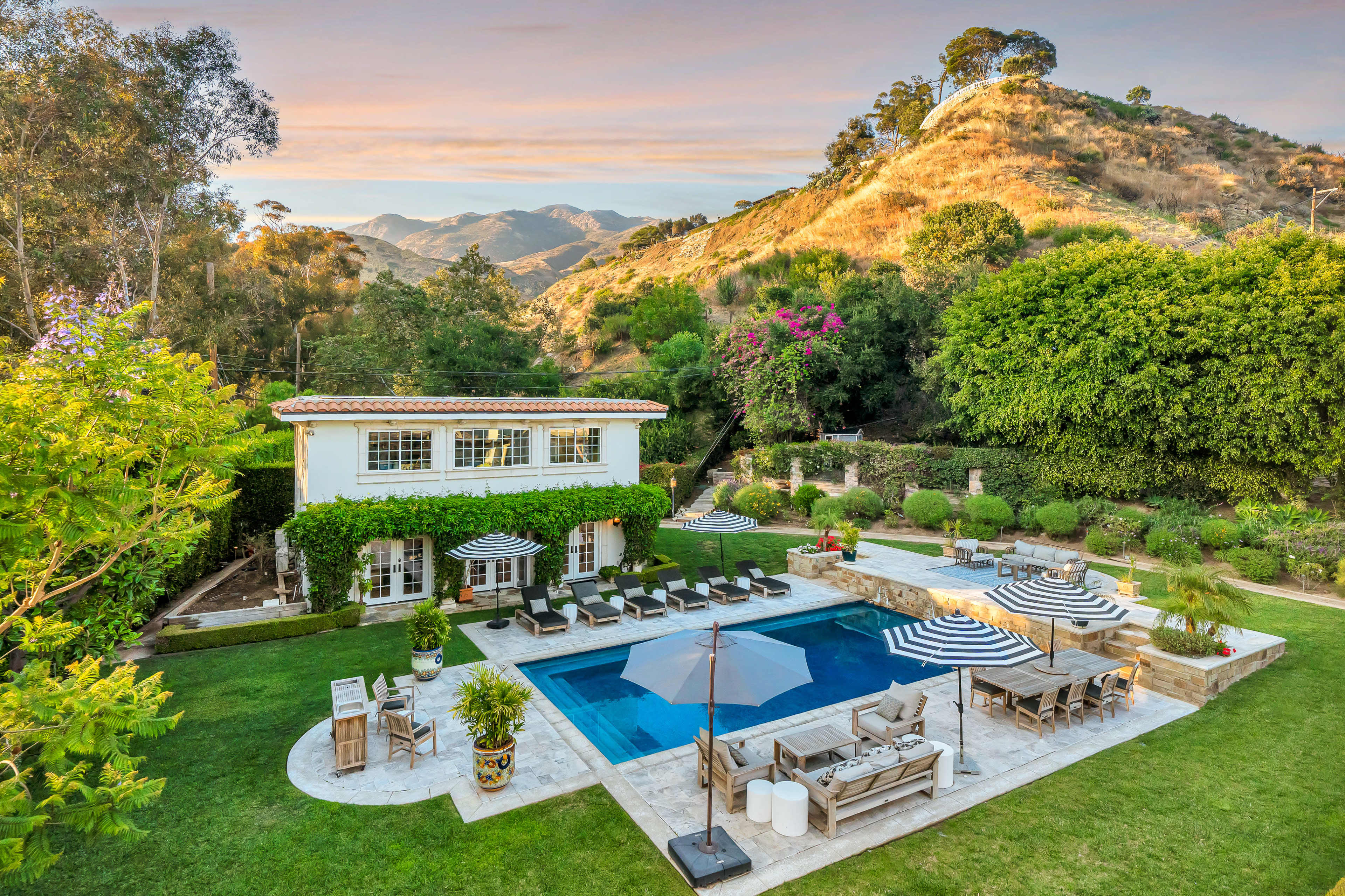 The image shows a spacious outdoor area of a house featuring a pool, lounge chairs, and surrounding greenery with hills in the background.