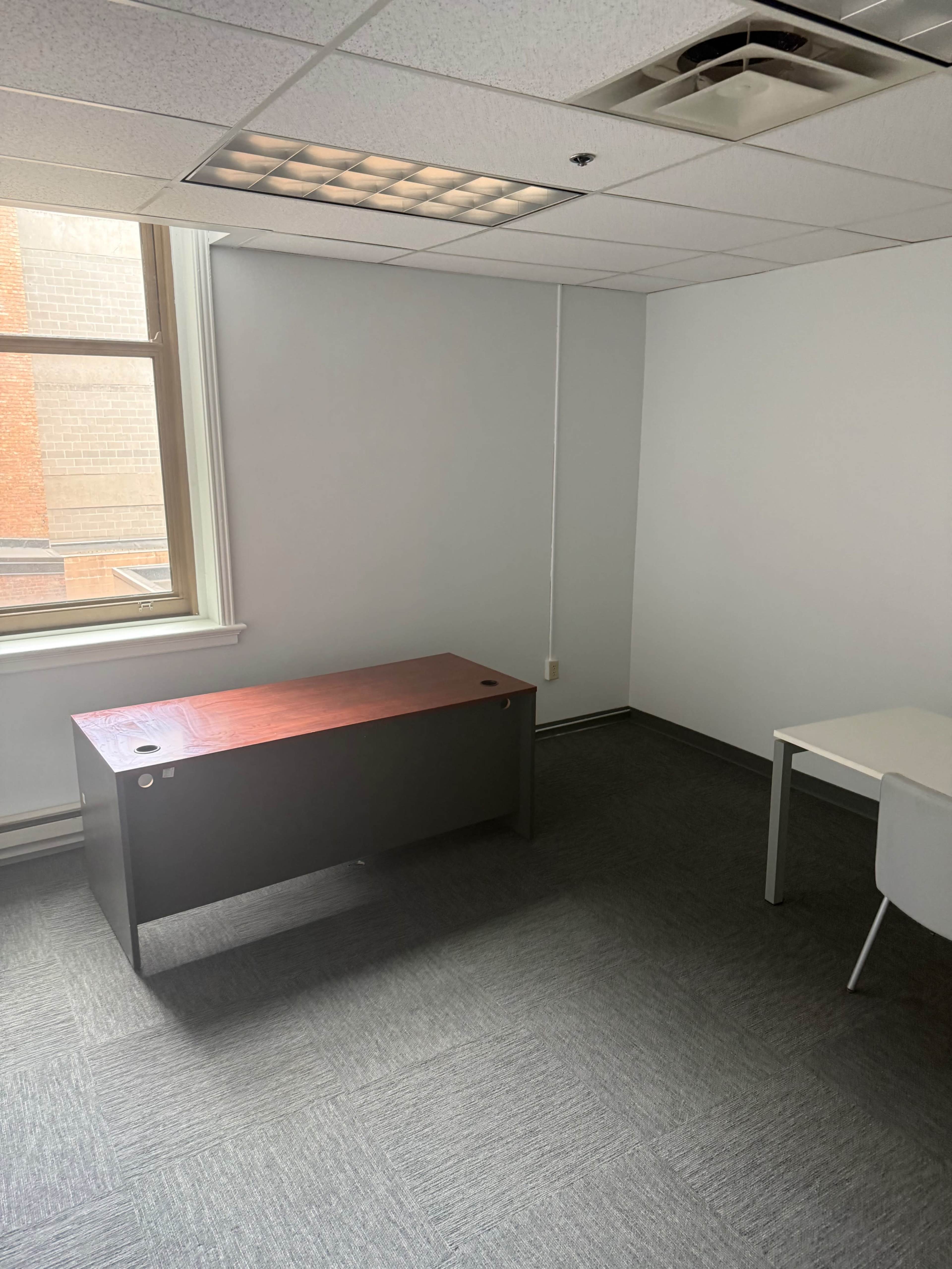 An empty office space features a dark partitioned desk and a small white table, with natural light coming through the window.