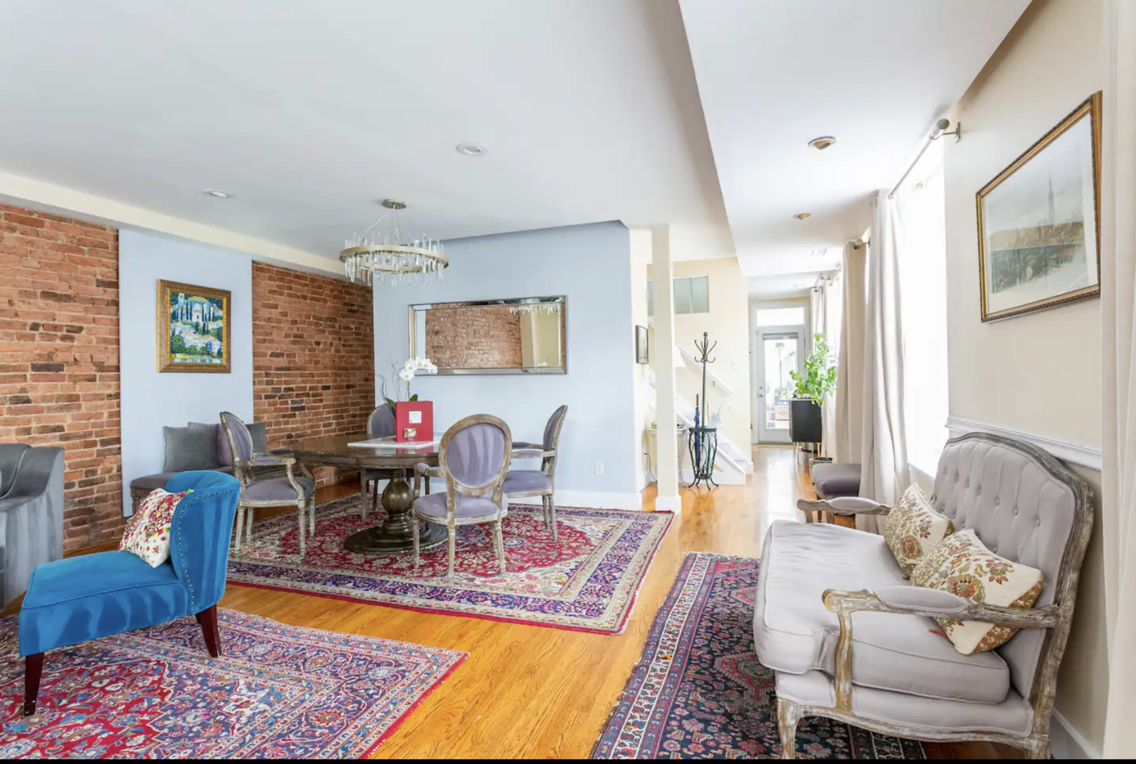 A spacious living area features a round dining table surrounded by chairs, a sofa, and exposed brick walls, complemented by area rugs and decorative accents.