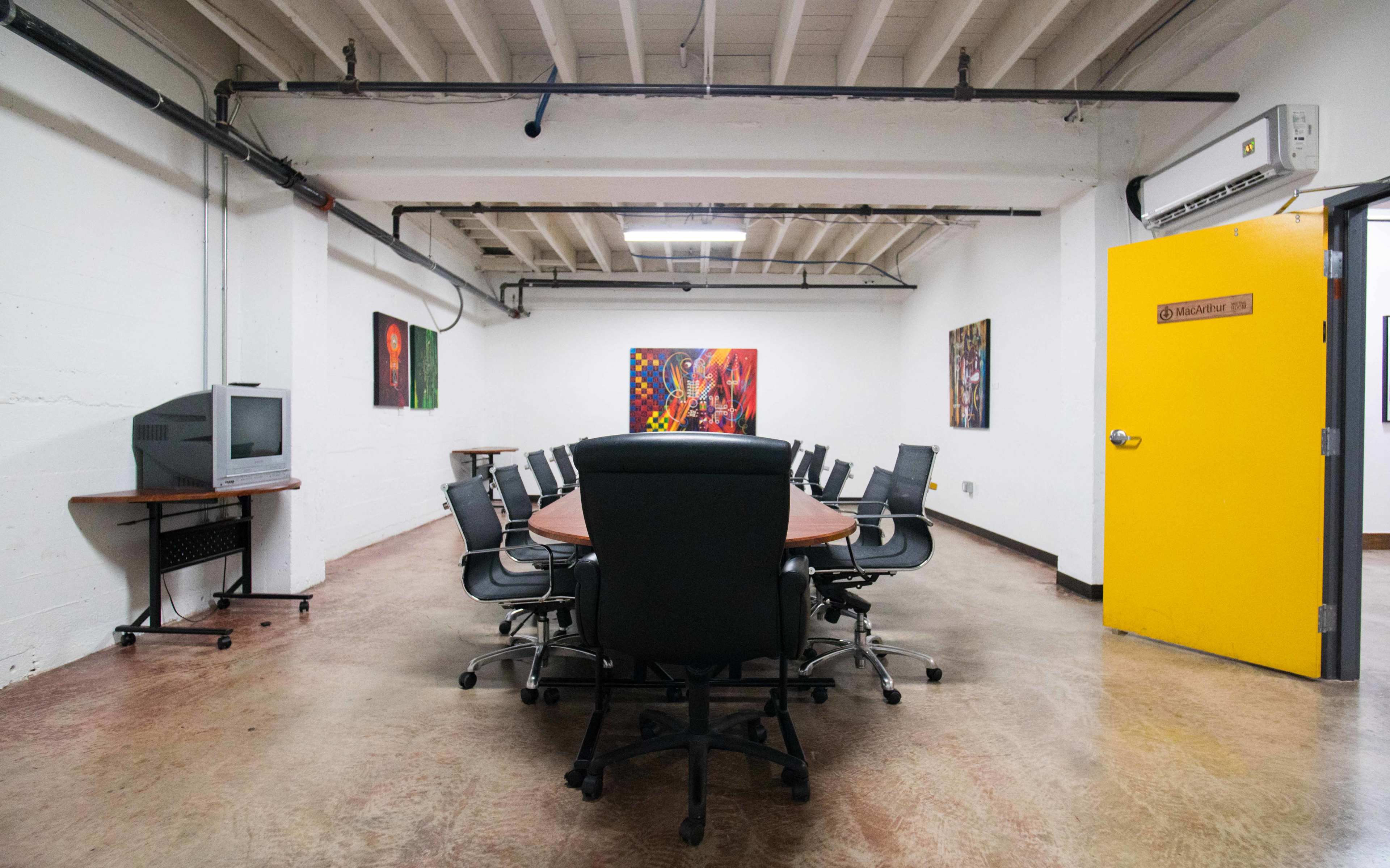 A large conference room features a rounded wooden table surrounded by black office chairs, with two abstract artworks displayed on the walls and a yellow door leading outside.