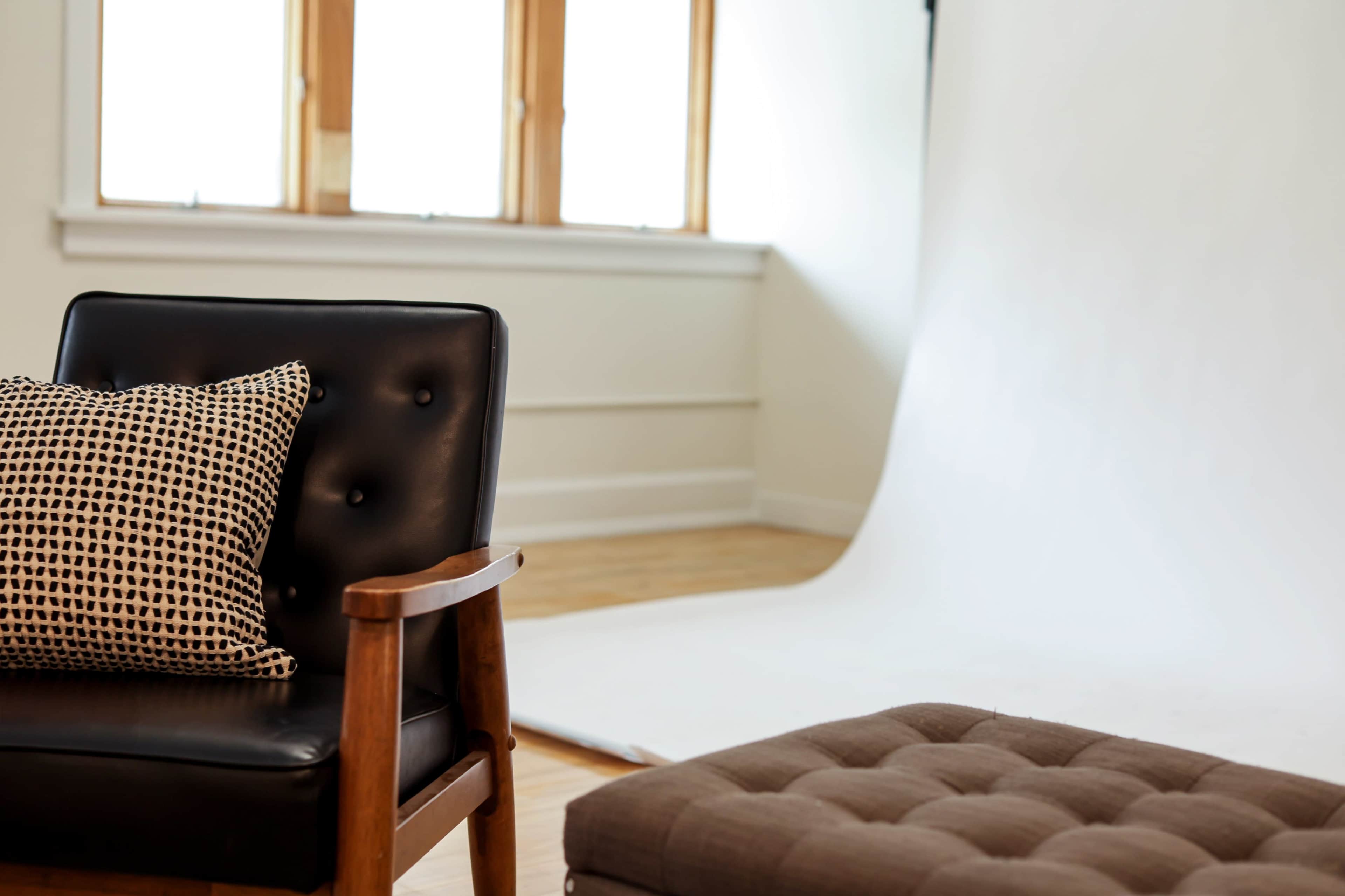 A black leather chair with a patterned cushion is positioned in a bright, minimalistic room featuring a white backdrop and large windows.