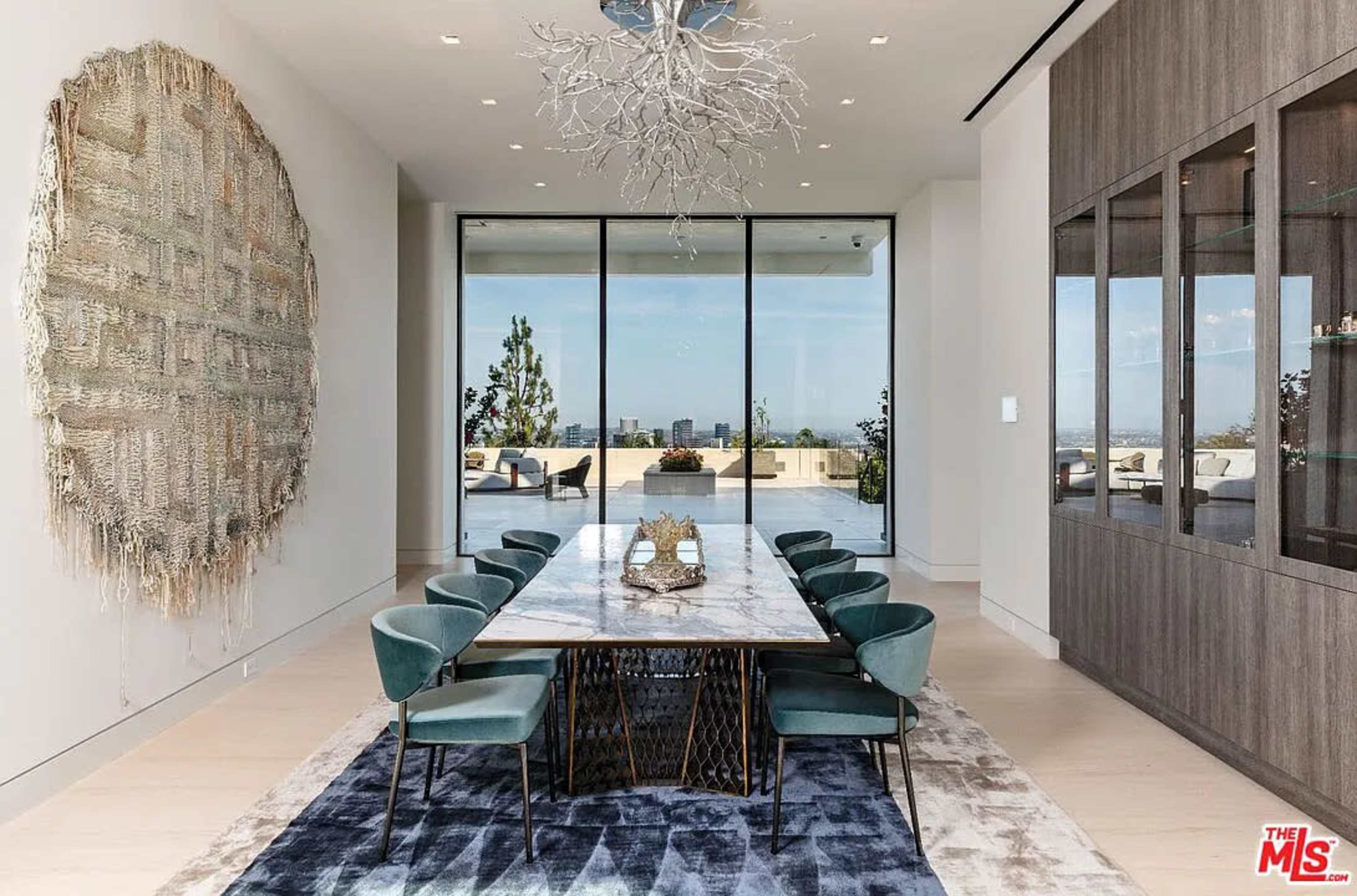 The image shows a modern dining room featuring a long marble table surrounded by teal chairs, with large windows offering a view of the outdoors and a circular textile wall hanging.