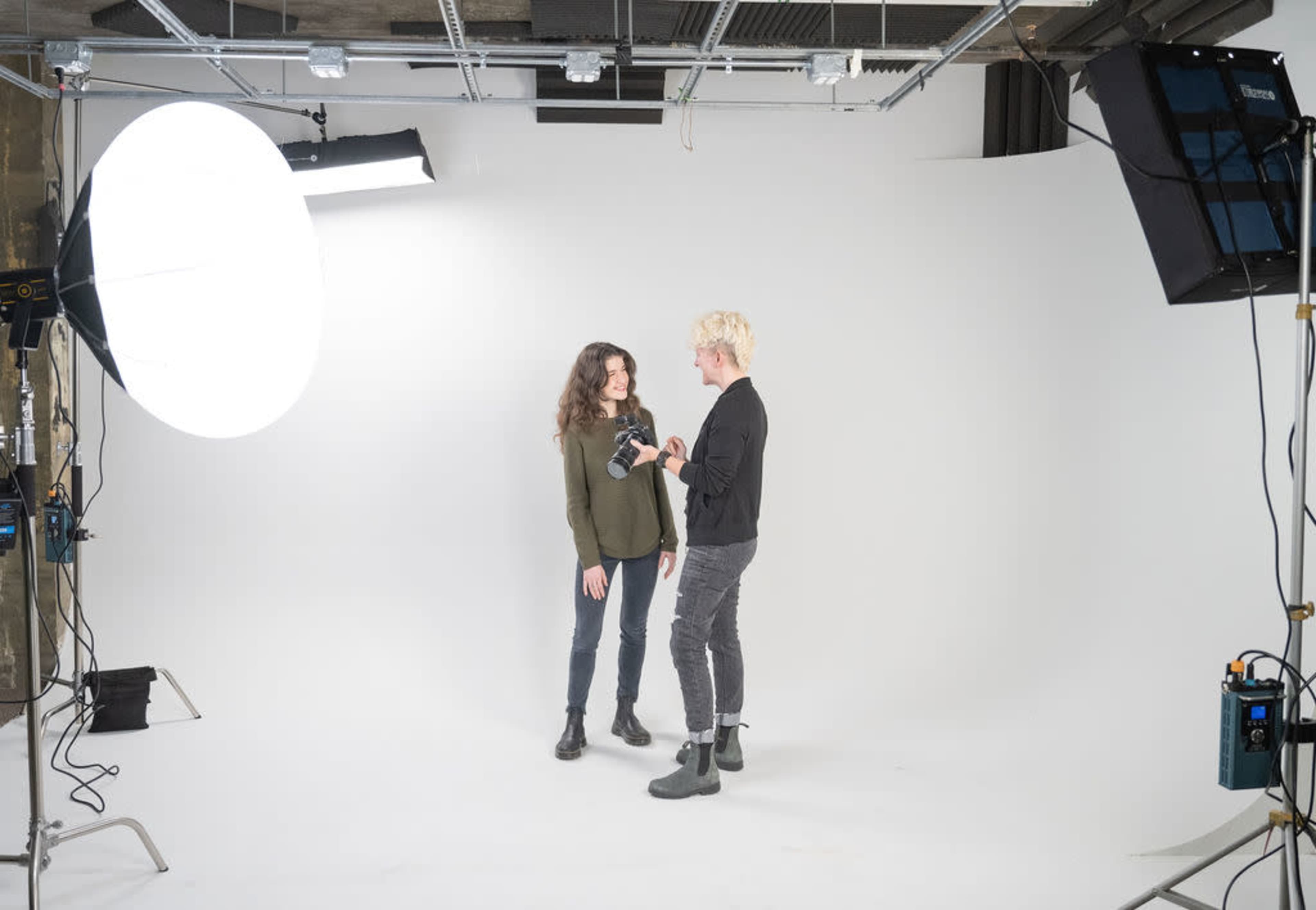 A photographer sets up lighting equipment while two models engage in conversation in a photo studio with a white backdrop.