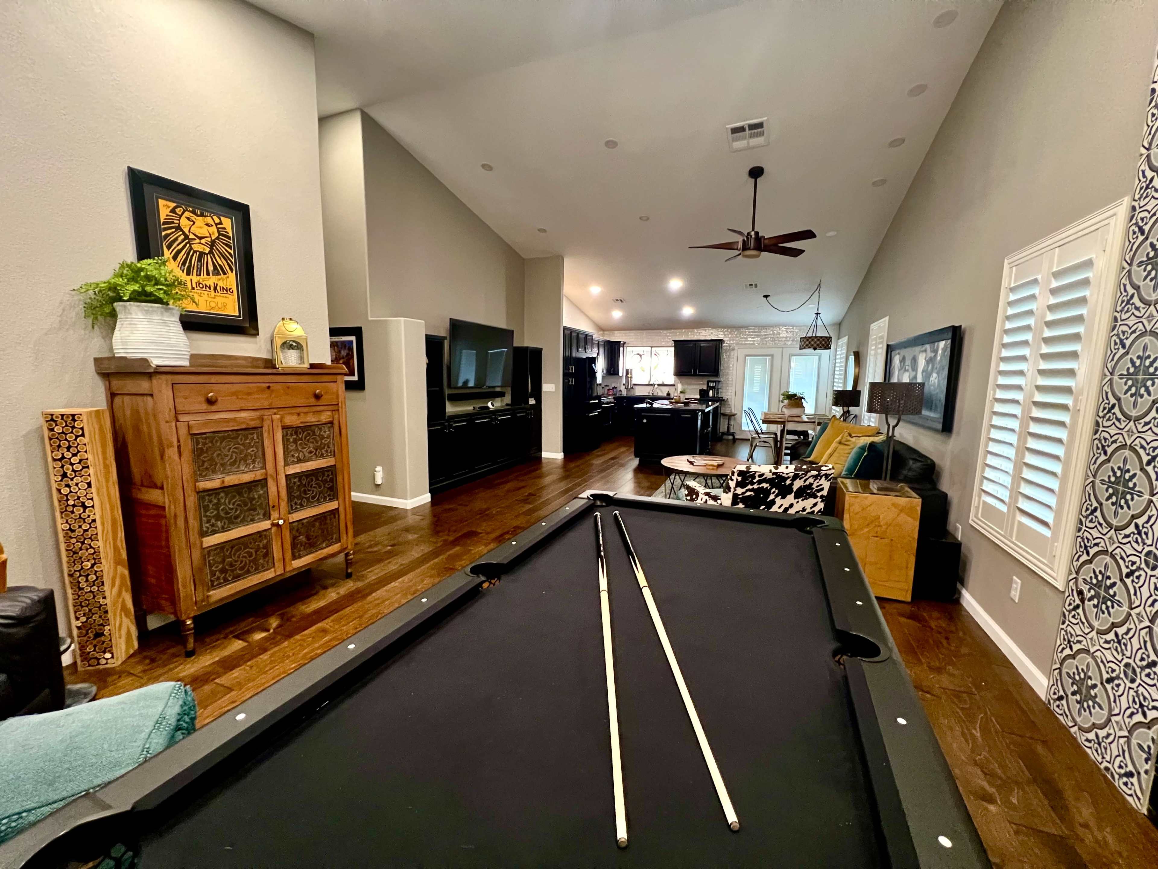 A spacious room features a pool table in the foreground, with a seating area and kitchen visible in the background.