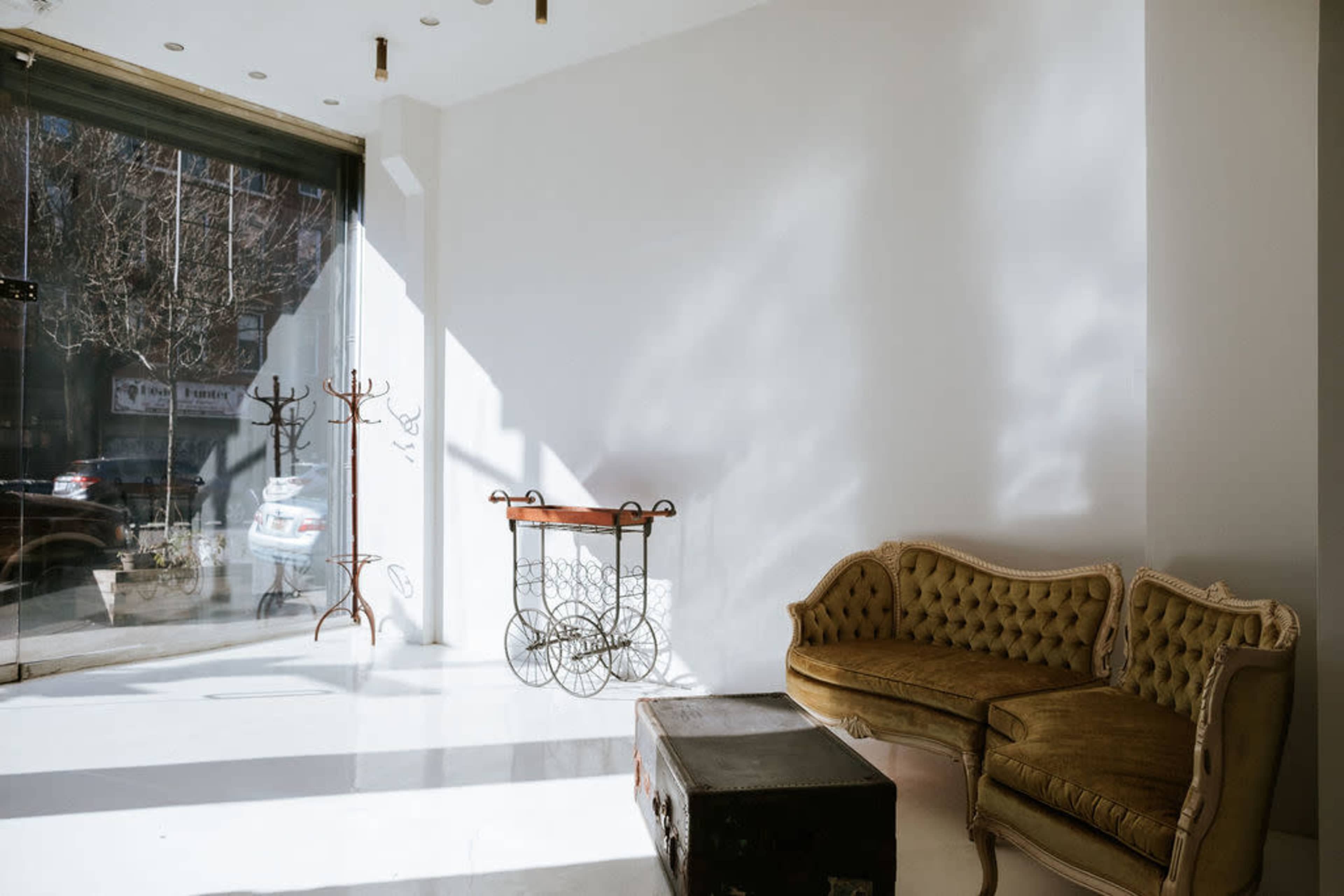 The scene features a brightly lit, minimalist interior with a vintage-style sofa, a metal cart, and a coat rack against a white wall.