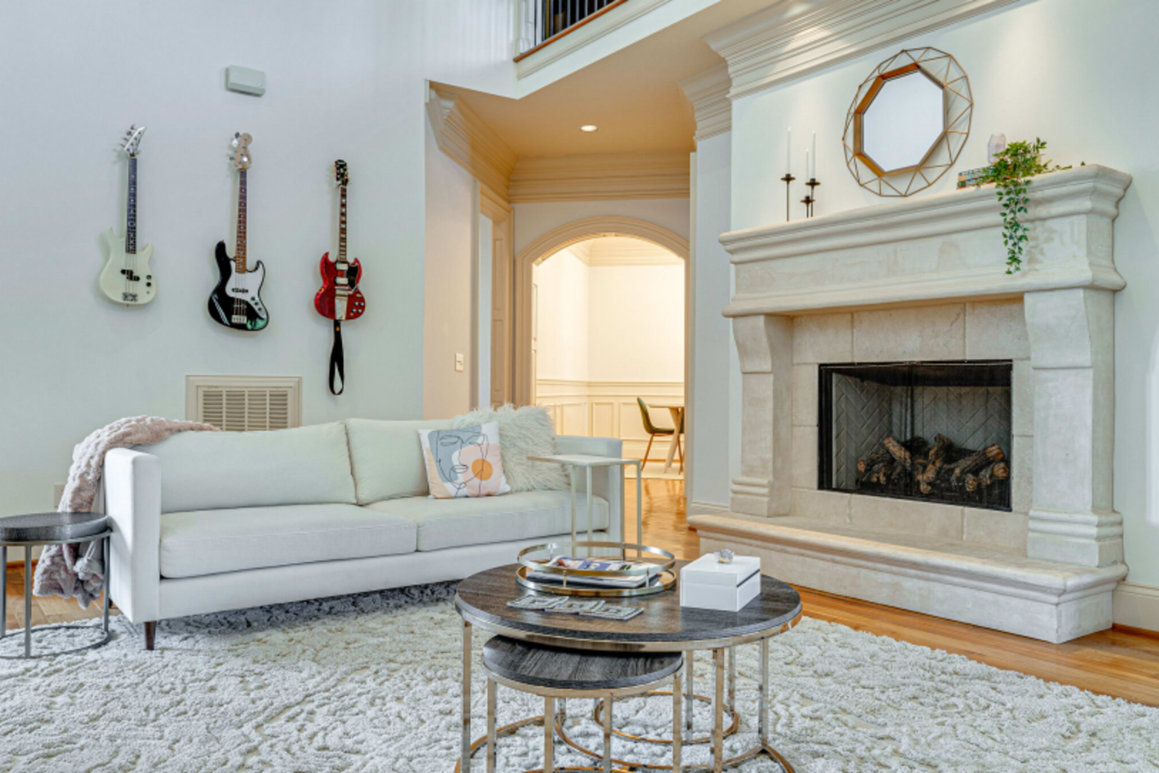 The image shows a living room featuring a white sofa, a stone fireplace, two coffee tables, and guitars hanging on the wall.