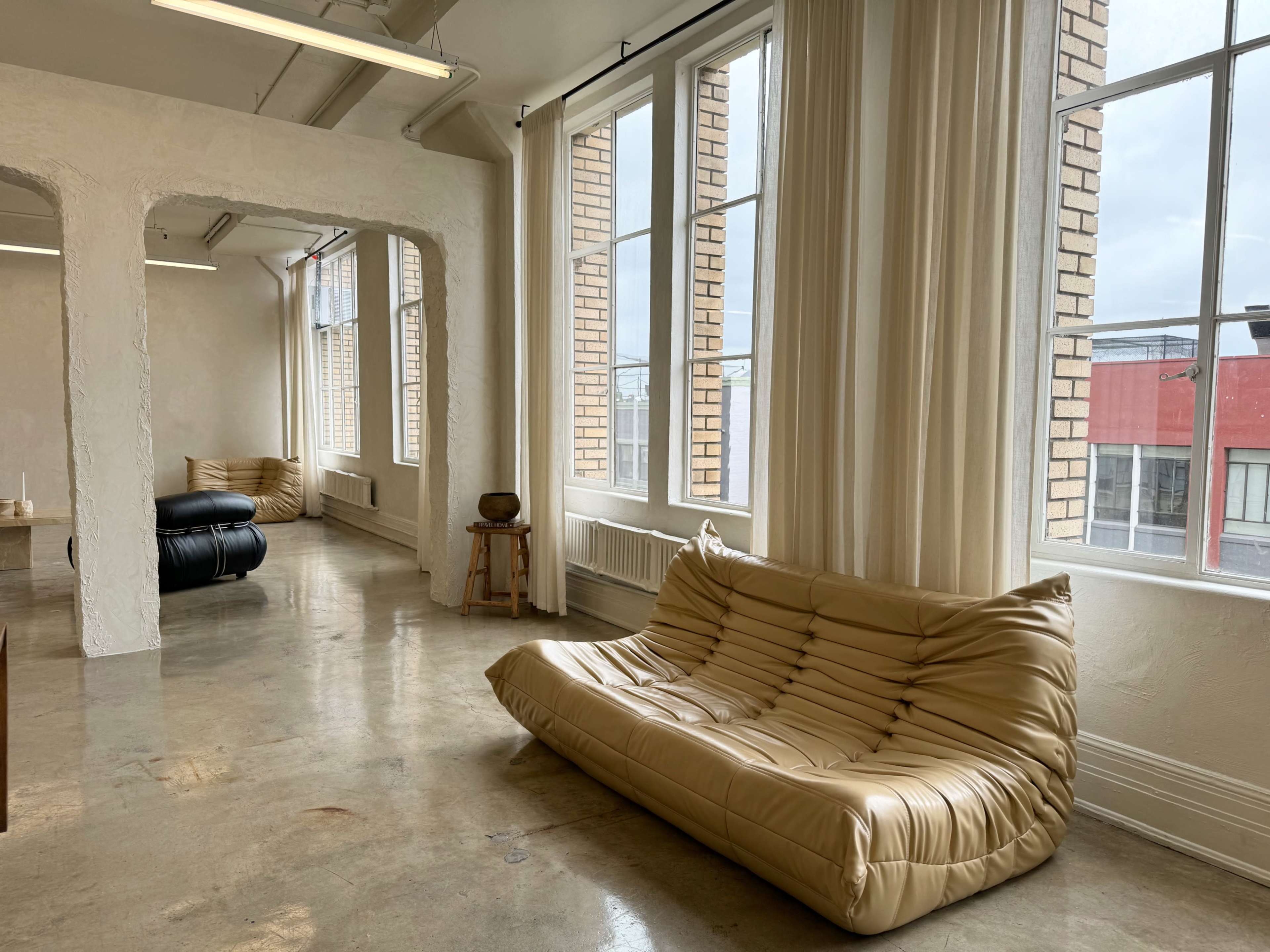The image shows a spacious interior with large windows, featuring a beige, sculptural sofa and a black sofa in a well-lit room with polished concrete flooring.