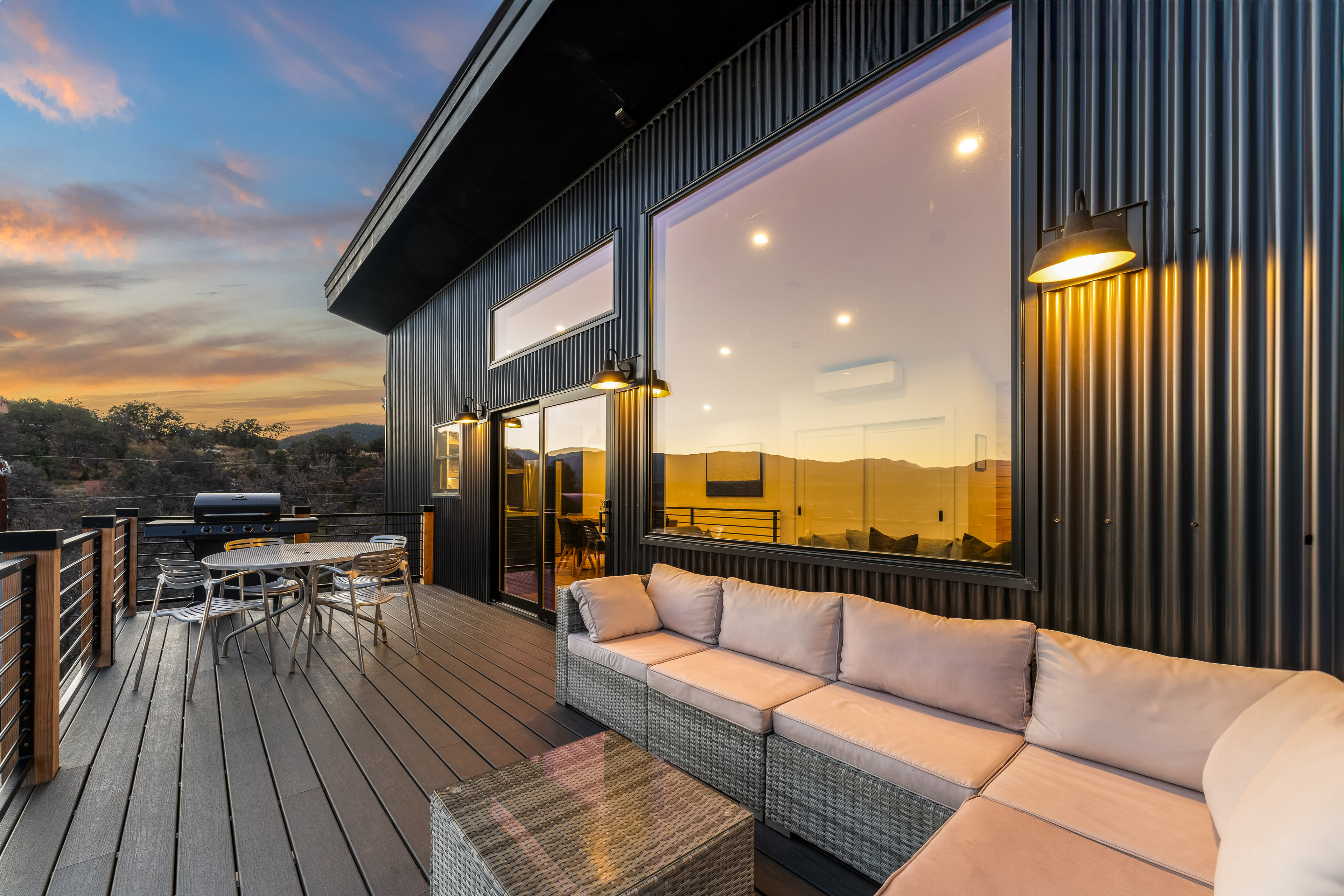A modern patio features a sectional sofa and a dining area, surrounded by large windows that reflect a sunset over the mountains.
