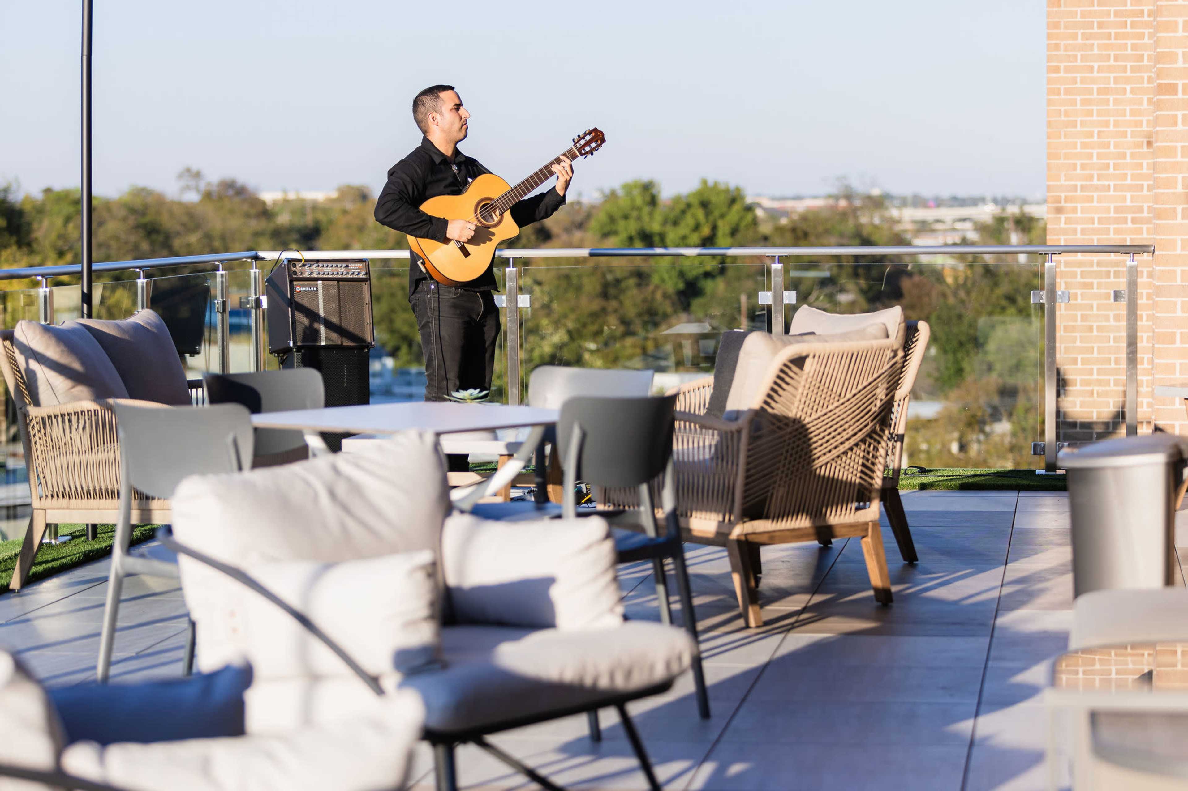 A musician plays a guitar on a terrace surrounded by outdoor furniture and greenery.