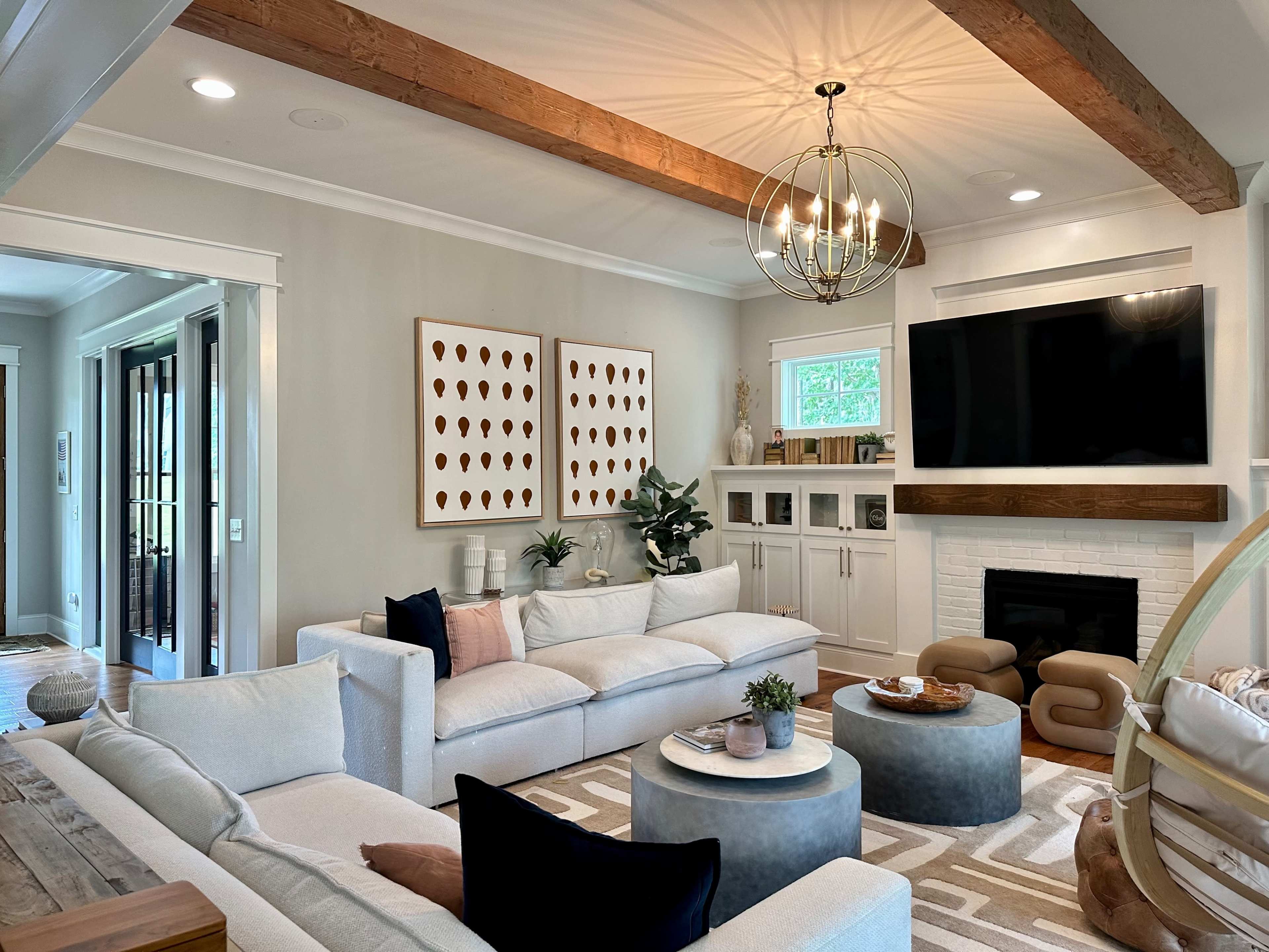 The living room features a light-colored sectional sofa, a round coffee table, and a mounted flat-screen TV above a white fireplace, with wooden beams and decorative artwork on the walls.
