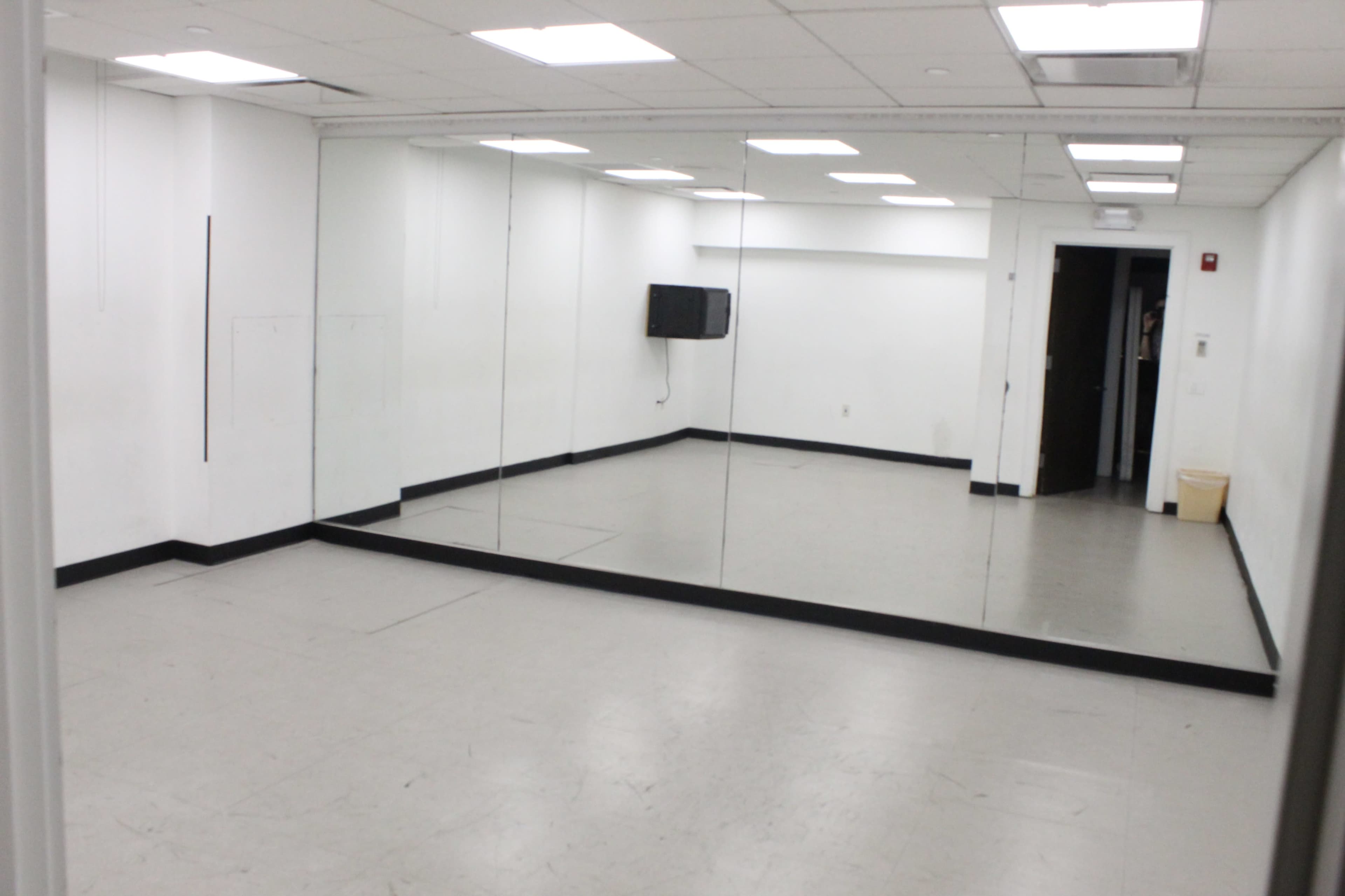 The image shows a sparsely furnished dance studio with mirrored walls and a small television mounted on one side.