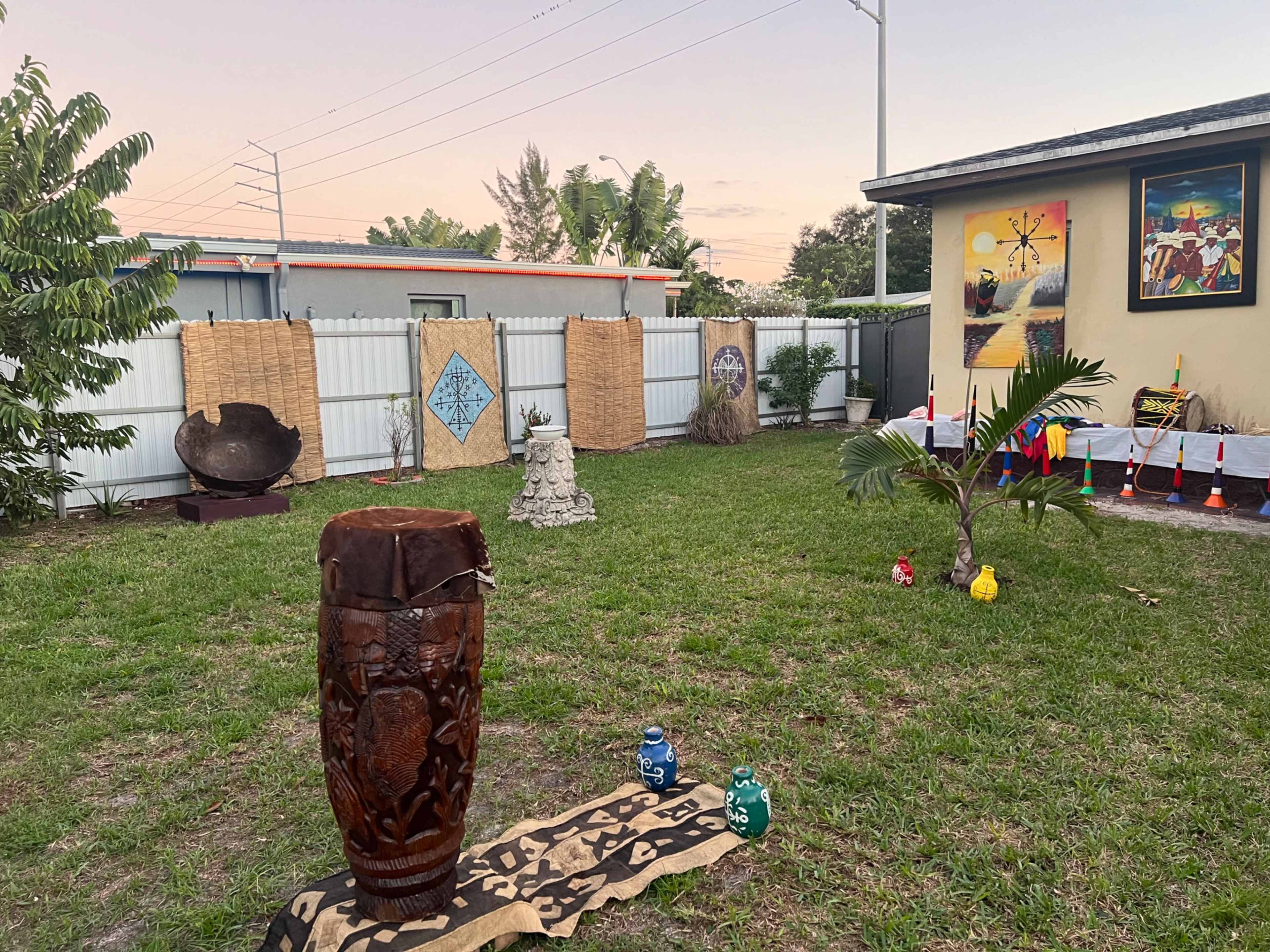 The scene features a grassy yard with various decorations, including a large carved drum, tribal patterns on the ground, and multiple art pieces arranged against a fence.