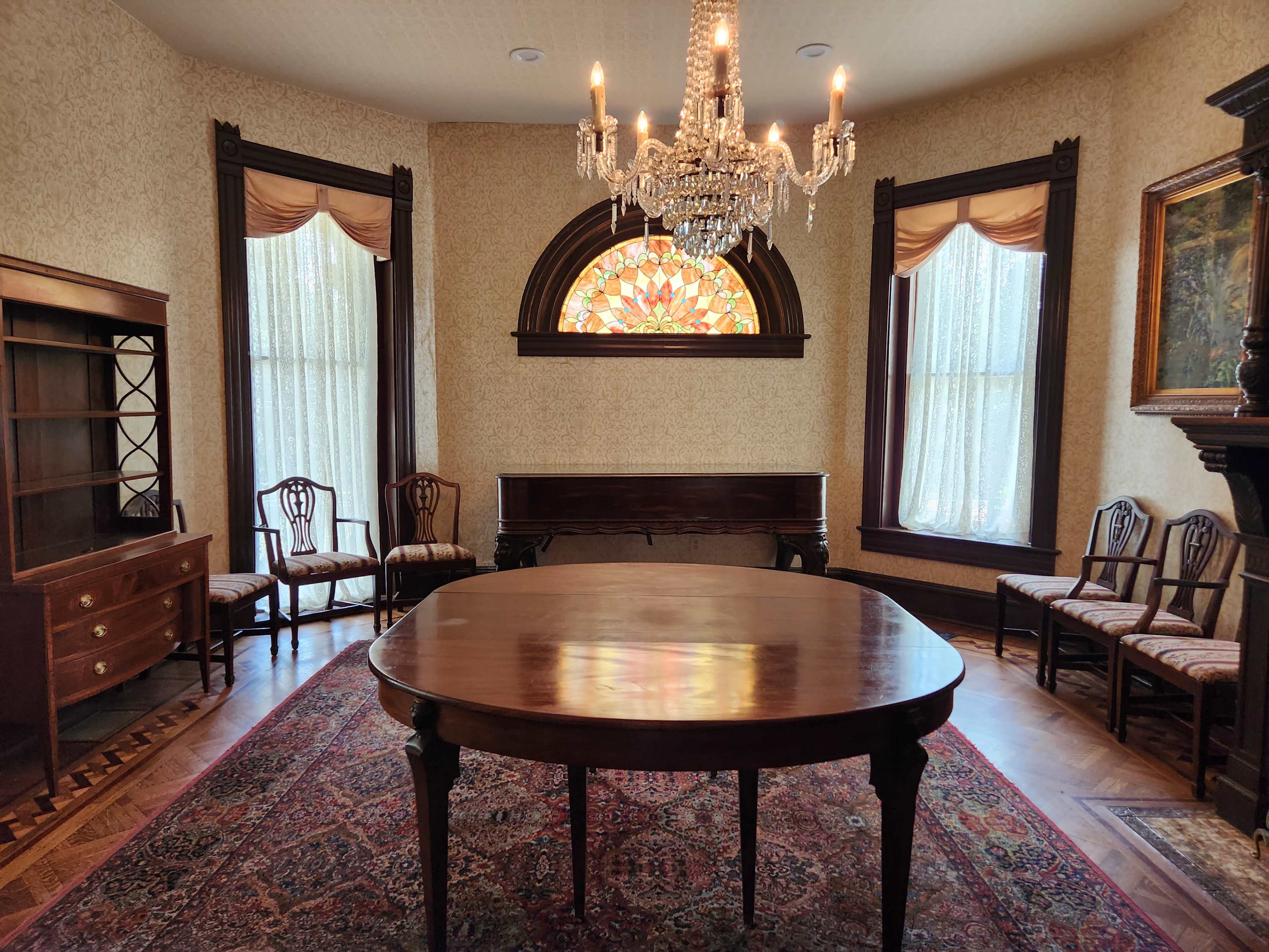 The image shows a well-lit room featuring a round wooden table, a stained glass window, a chandelier, and antique seating arranged around the space.