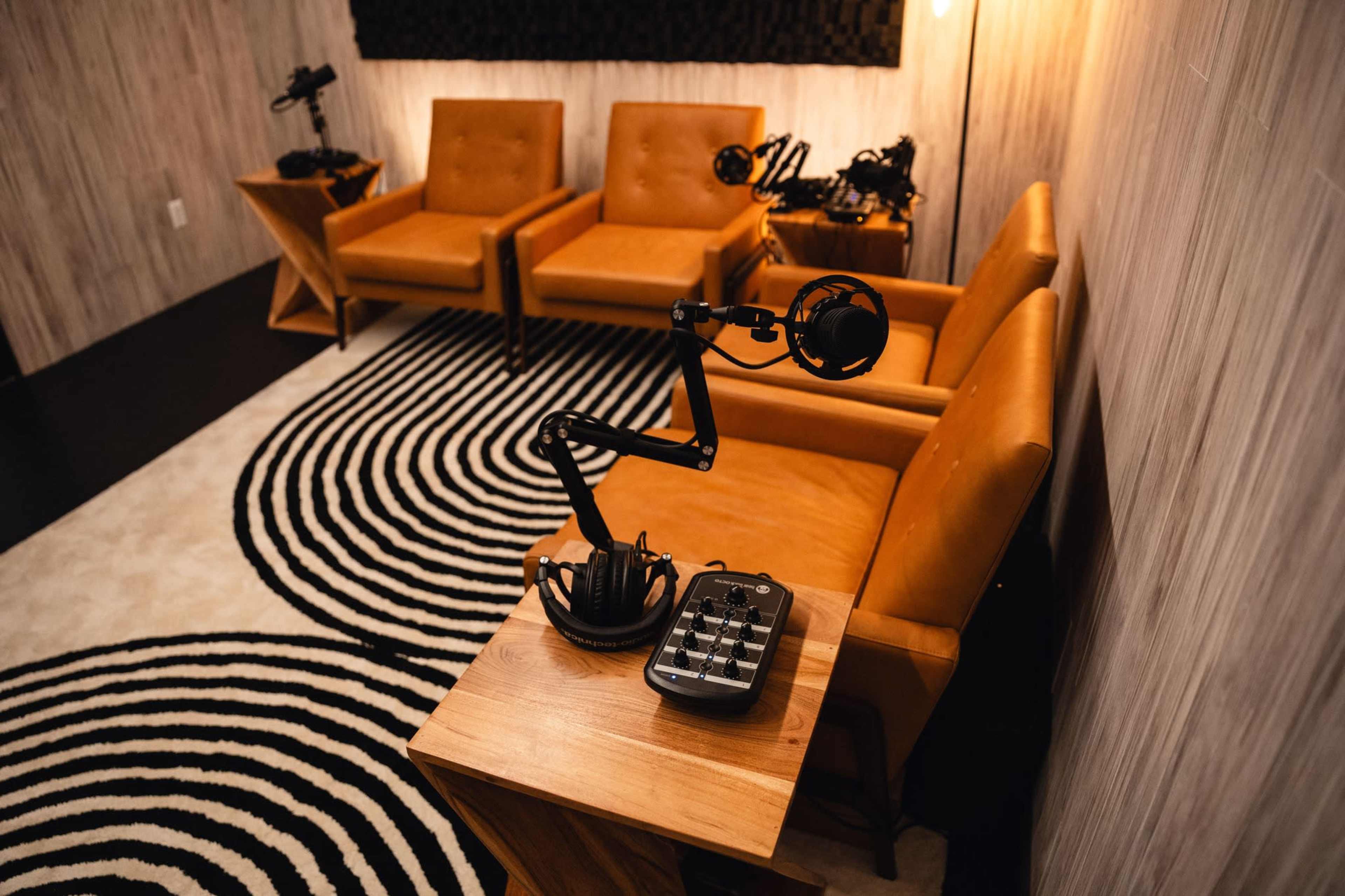 The image shows a small recording studio with two brown chairs, a microphone setup, and a mixing console on a wooden table, all situated on a black and white striped rug.