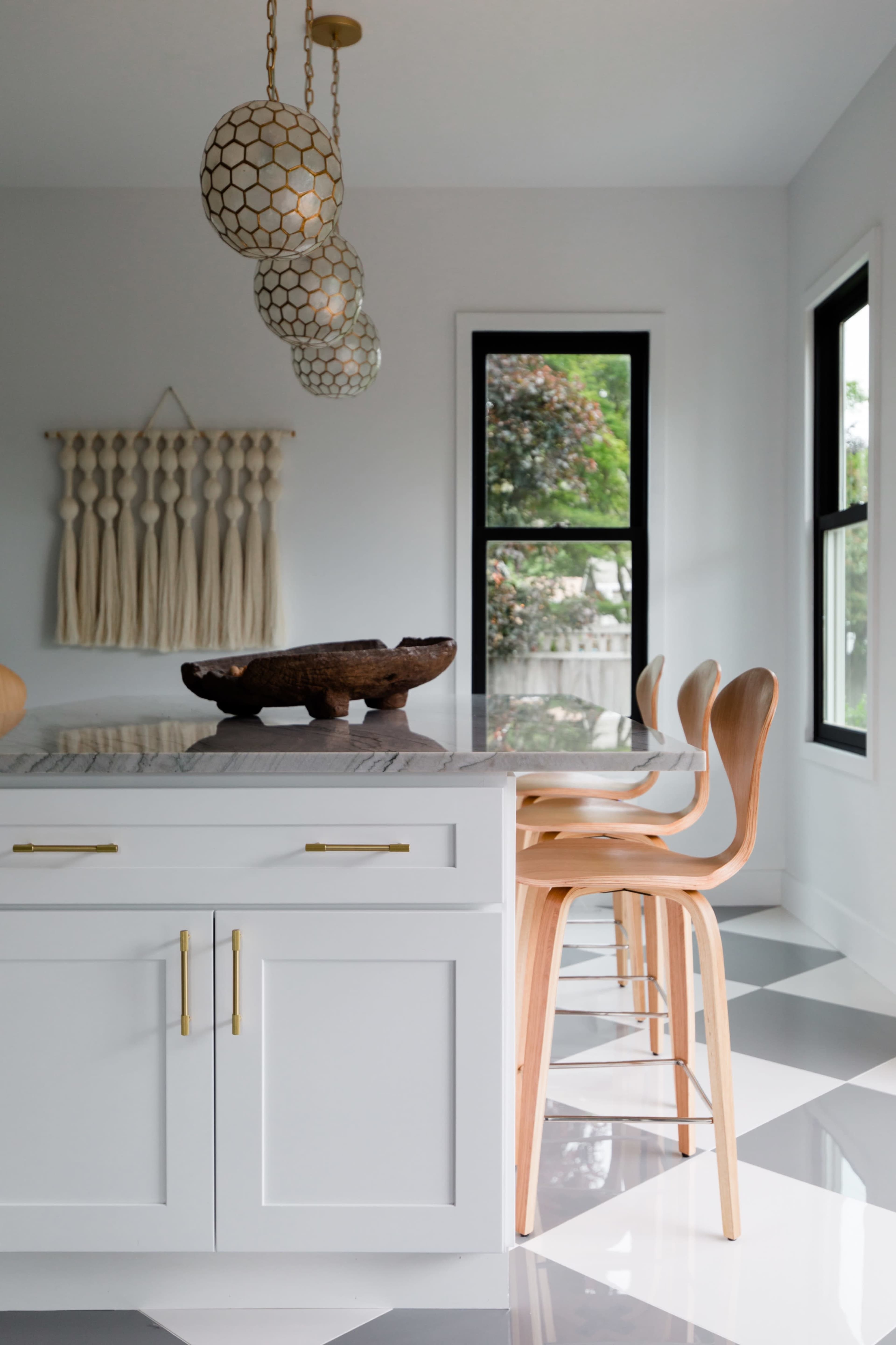 A modern kitchen features a spacious island with a decorative bowl and wooden stools, complemented by pendant lighting and large windows.