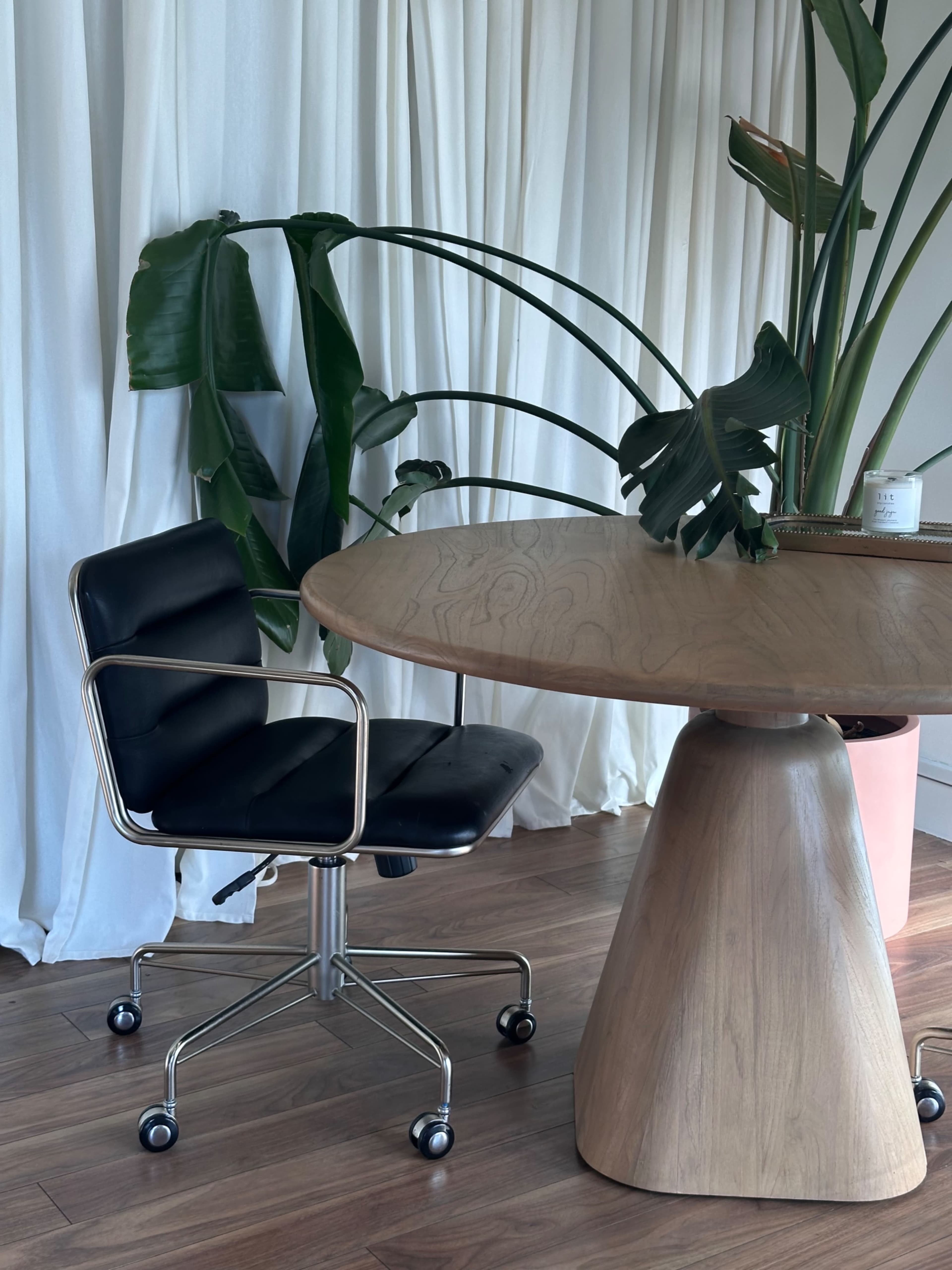 A black swivel chair sits next to a round wooden table in a room with white drapes and potted plants.