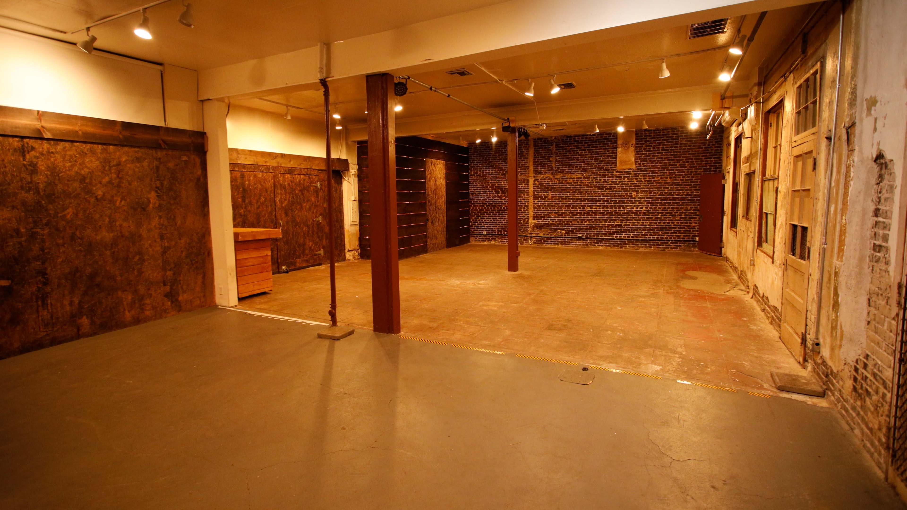 An empty industrial-style interior space with exposed brick walls and a concrete floor, featuring wooden accents and overhead lighting.