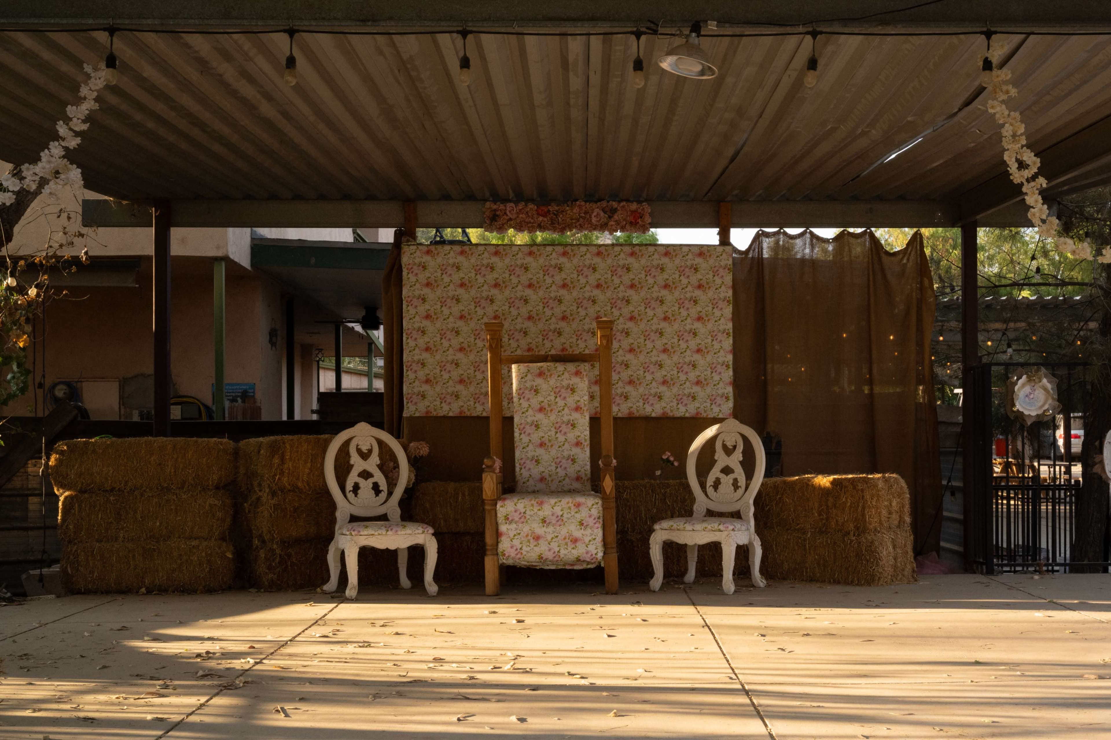 The image shows a decorated stage area with a floral backdrop, a large throne chair in the center, and two white chairs on either side, all positioned in front of a rustic setting with hay bales.