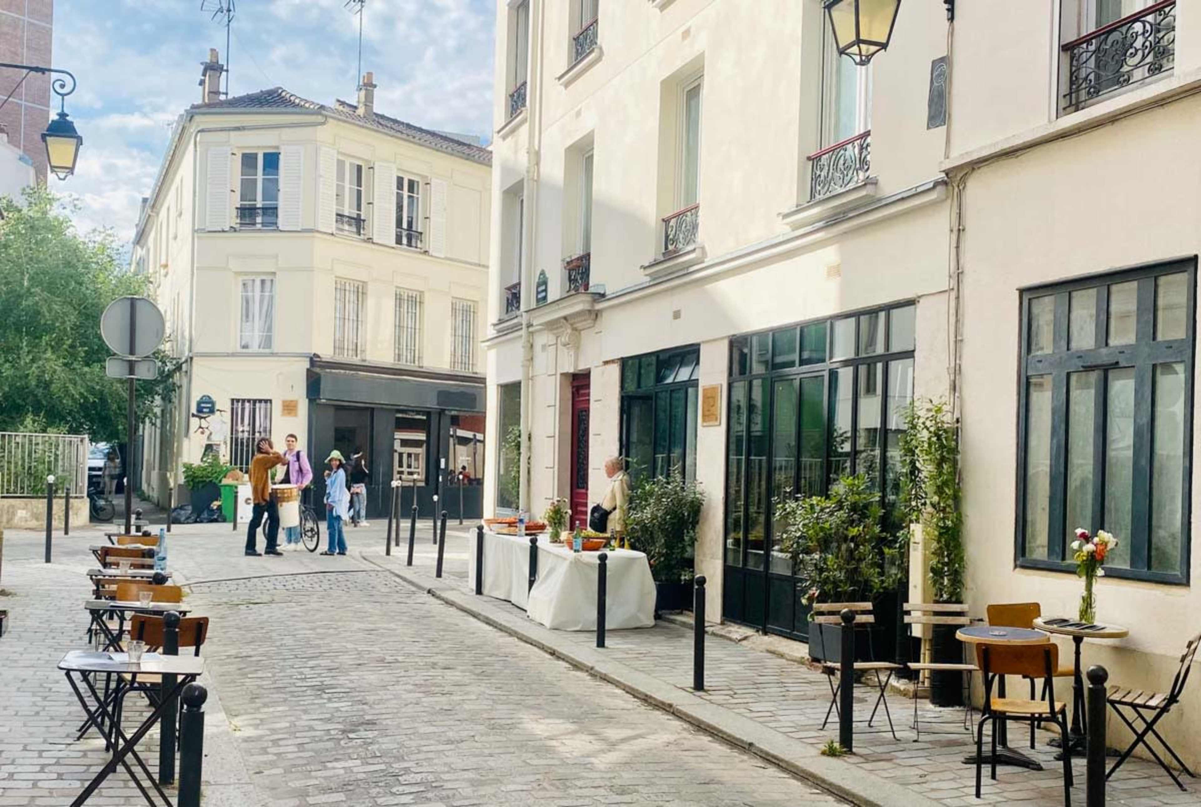 A cobblestone street is lined with small tables and chairs outside a café, with people gathered and a decorative table set up nearby.