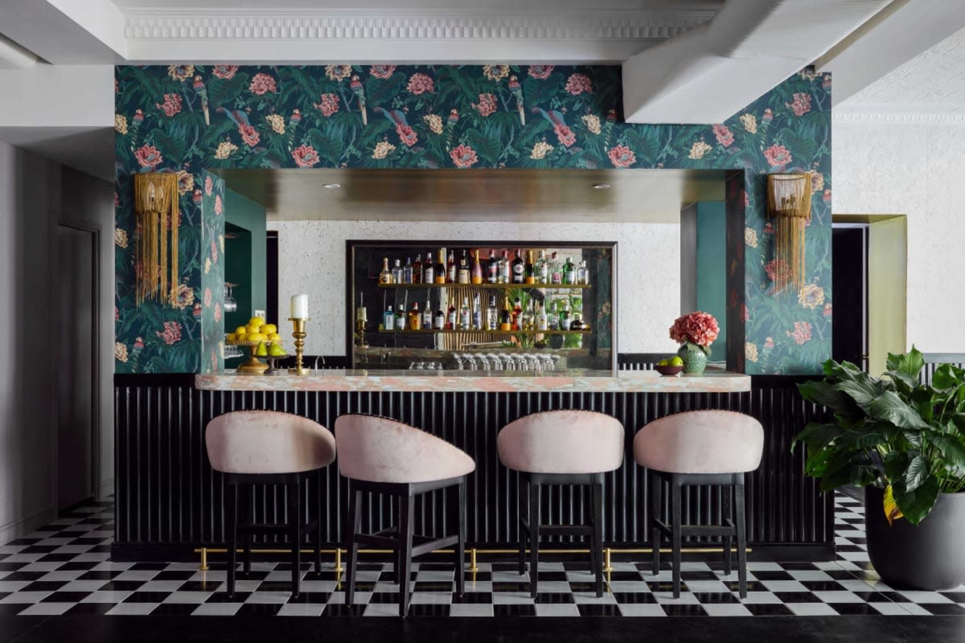 A stylish bar area featuring a floral-patterned wallpaper, a marble countertop, and four pink upholstered stools arranged in front of a well-stocked liquor display.