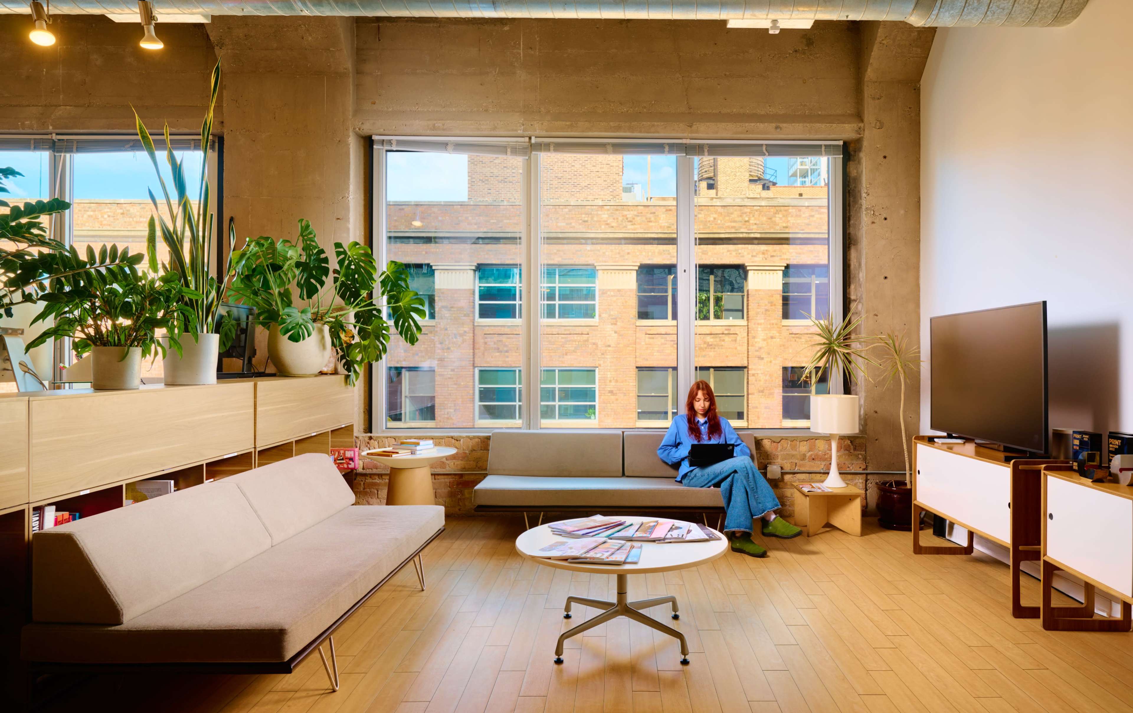A woman sits in a modern living space with large windows, plants, and minimalistic furniture.