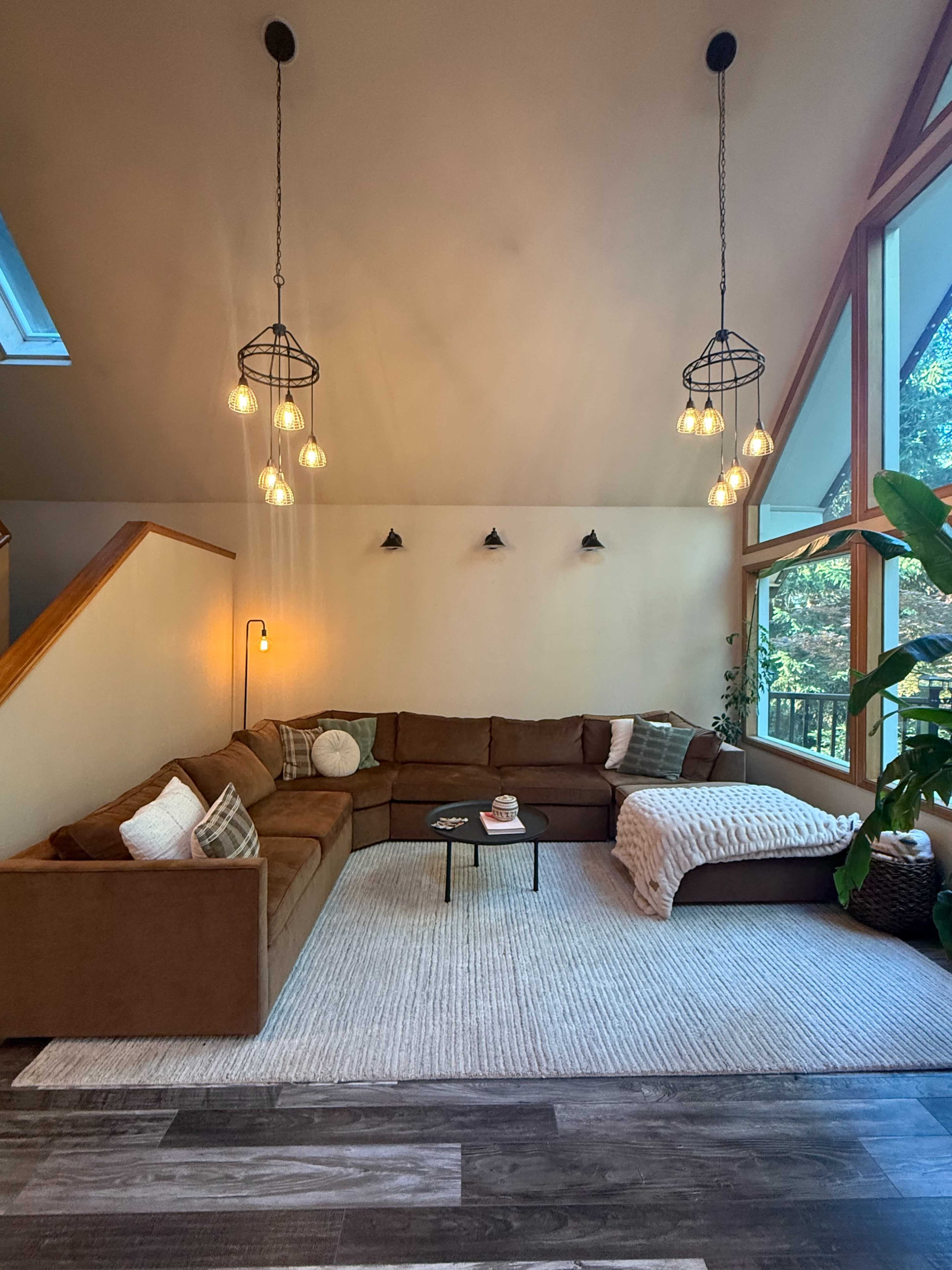 The image shows a modern living room with a large brown sectional sofa, a round coffee table, and several hanging light fixtures against a backdrop of large windows.
