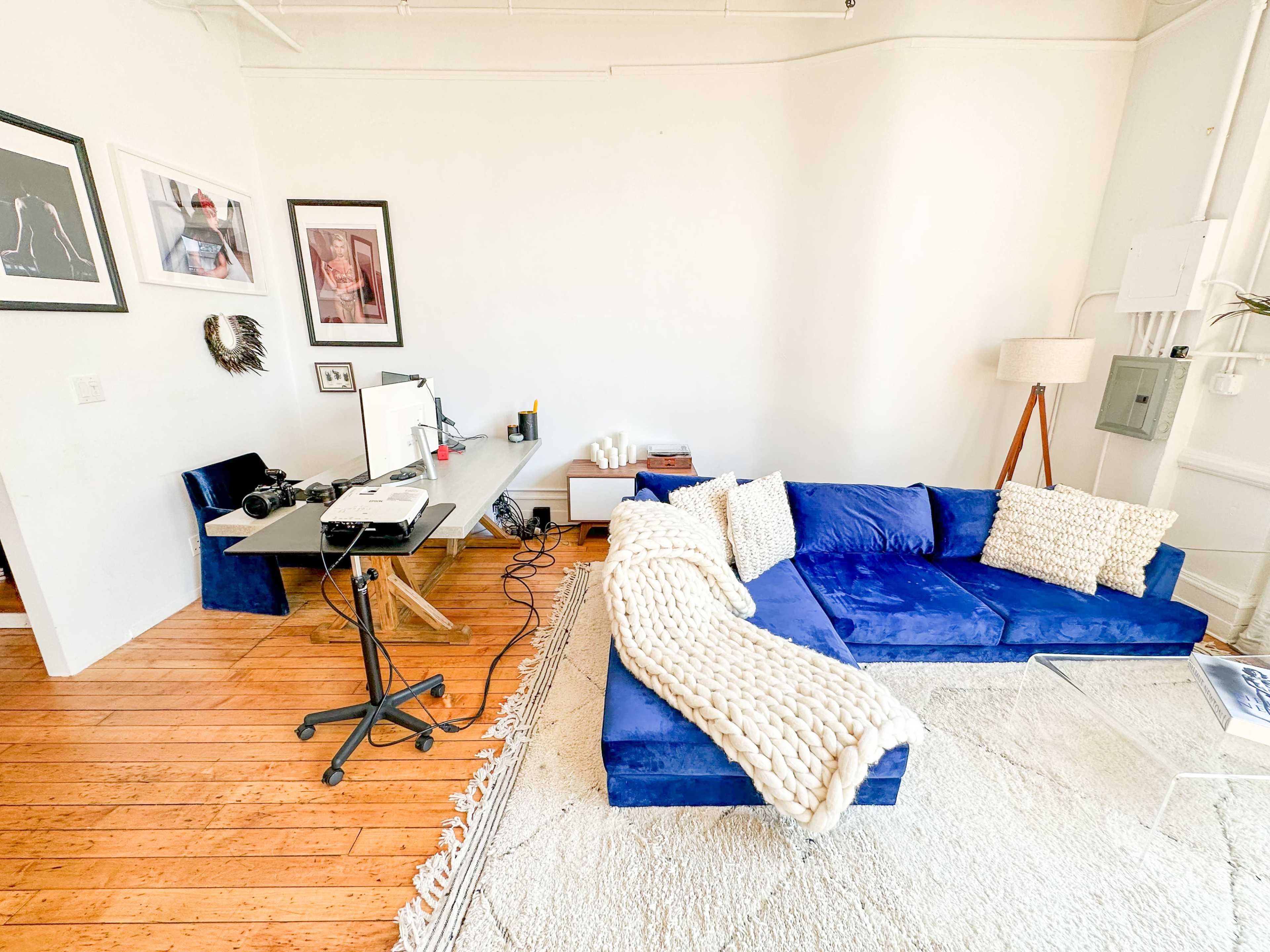 A modern living space with a blue sectional sofa, a small desk set up with a computer, and several framed artworks on the walls.
