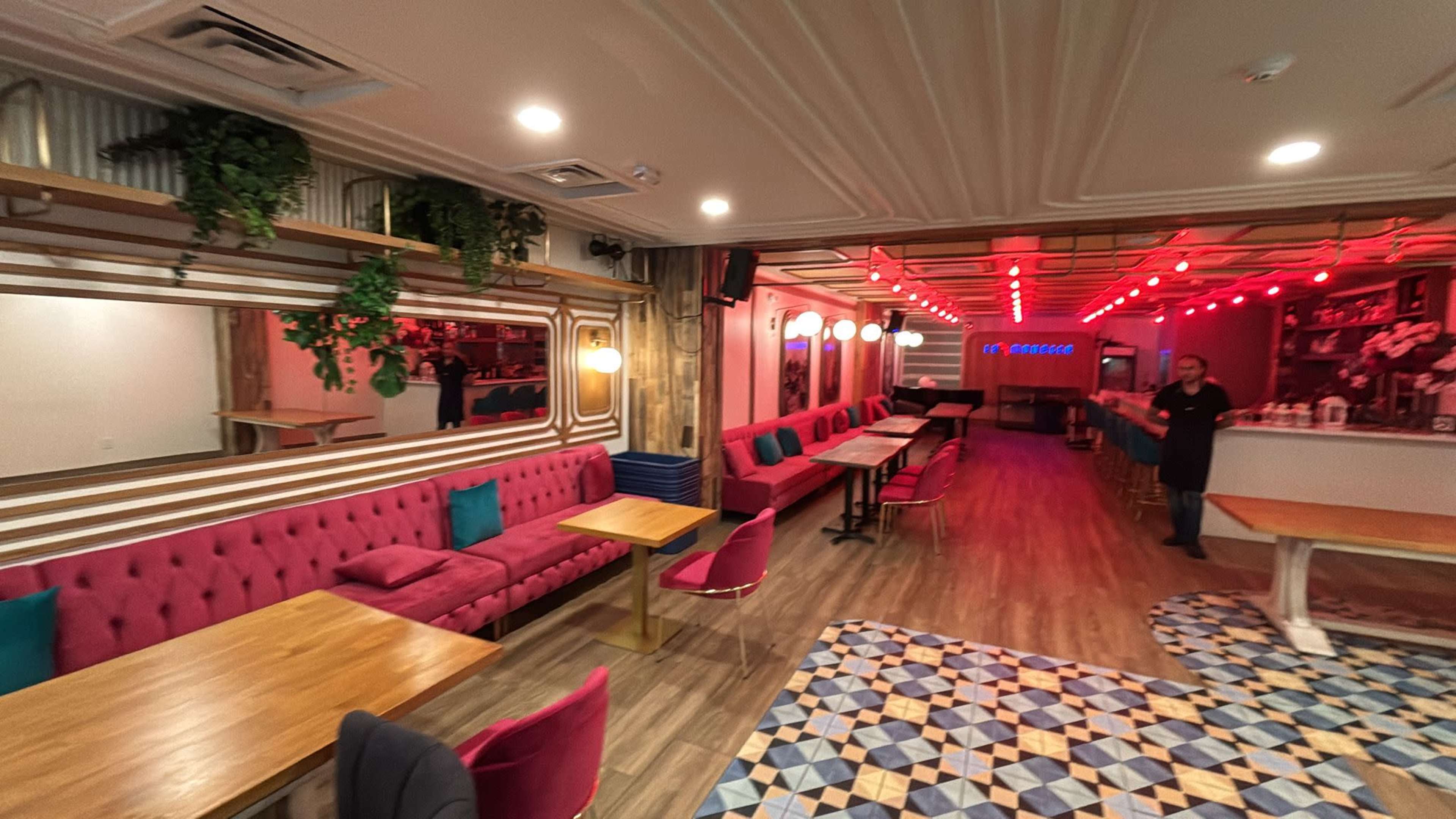The image shows a modern lounge or restaurant with pink upholstered seating, wooden tables, and vibrant lighting.