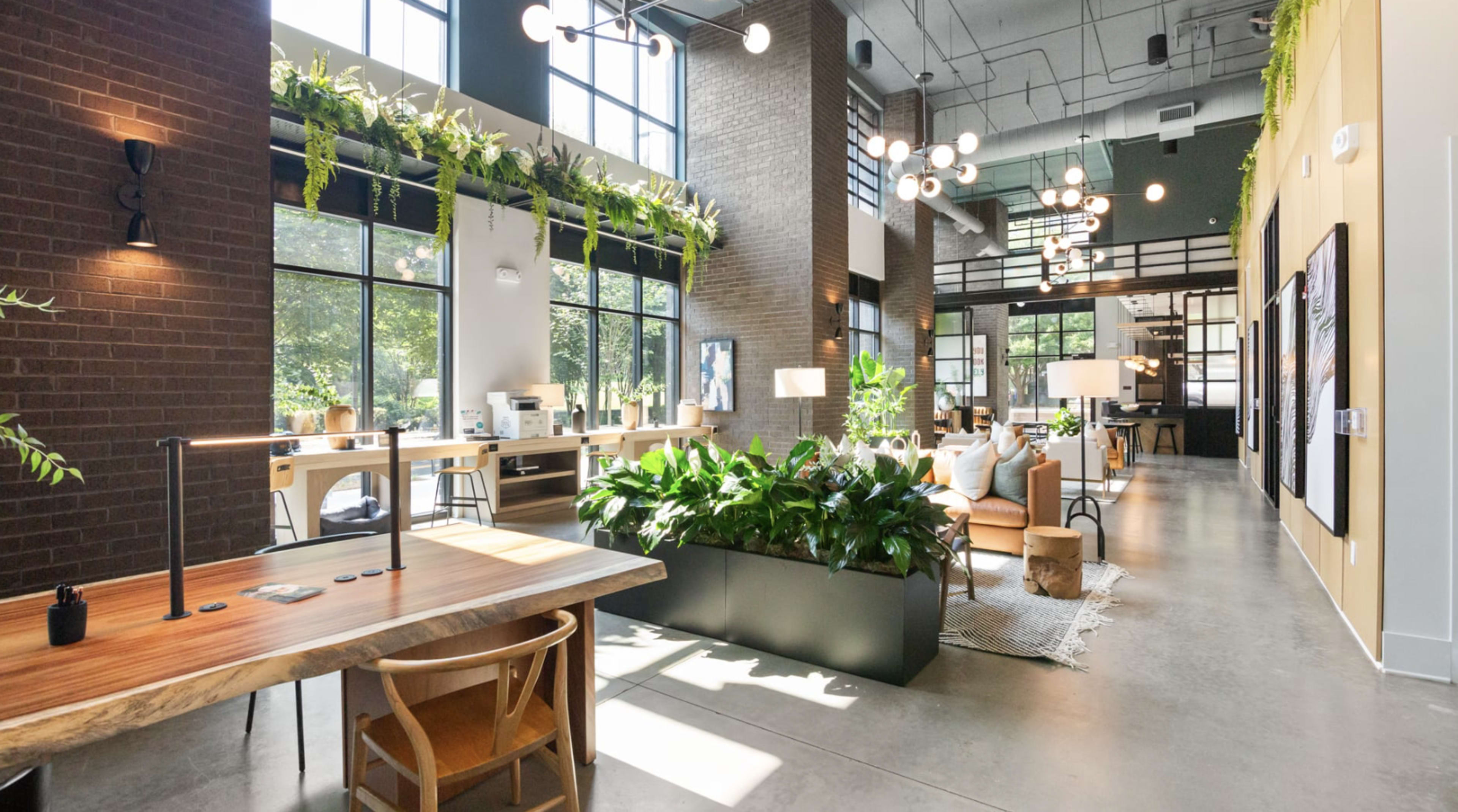 The image shows a modern interior of an open-plan office space with large windows, greenery, and contemporary furniture arranged for work and relaxation.