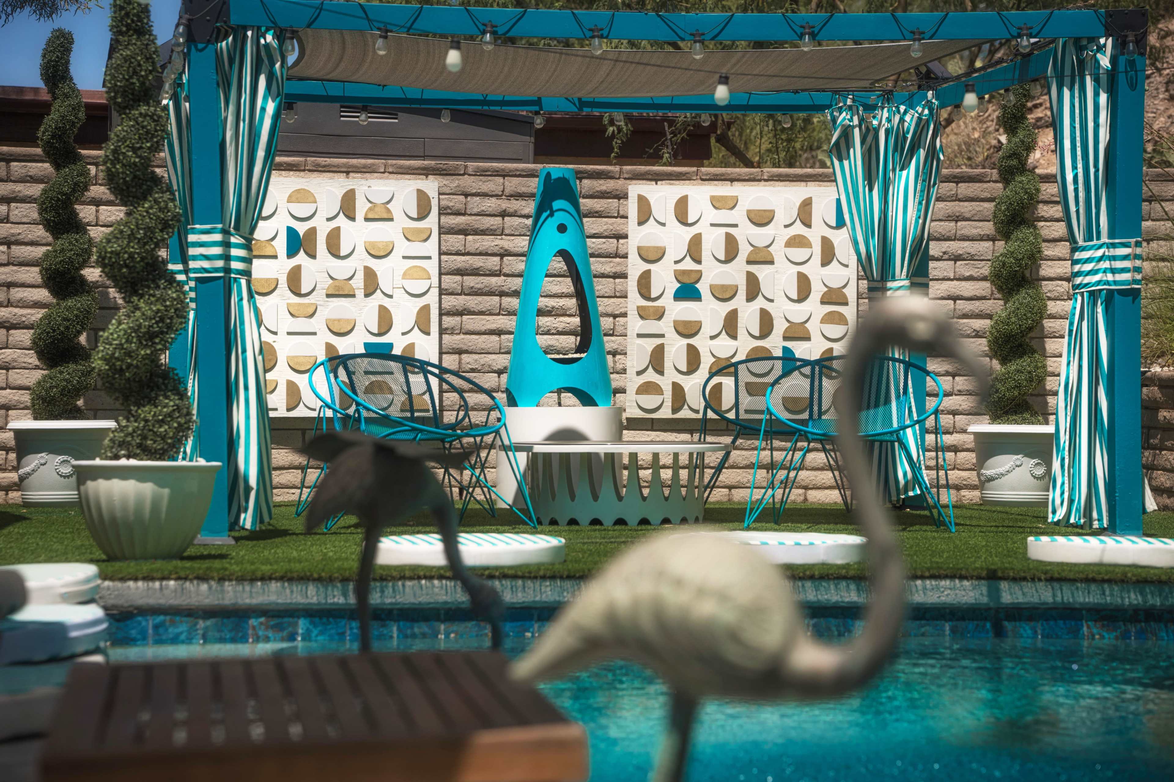 A backyard pool area features a turquoise canopy with seating, decorative topiaries, and flamingo sculptures beside a clear pool.