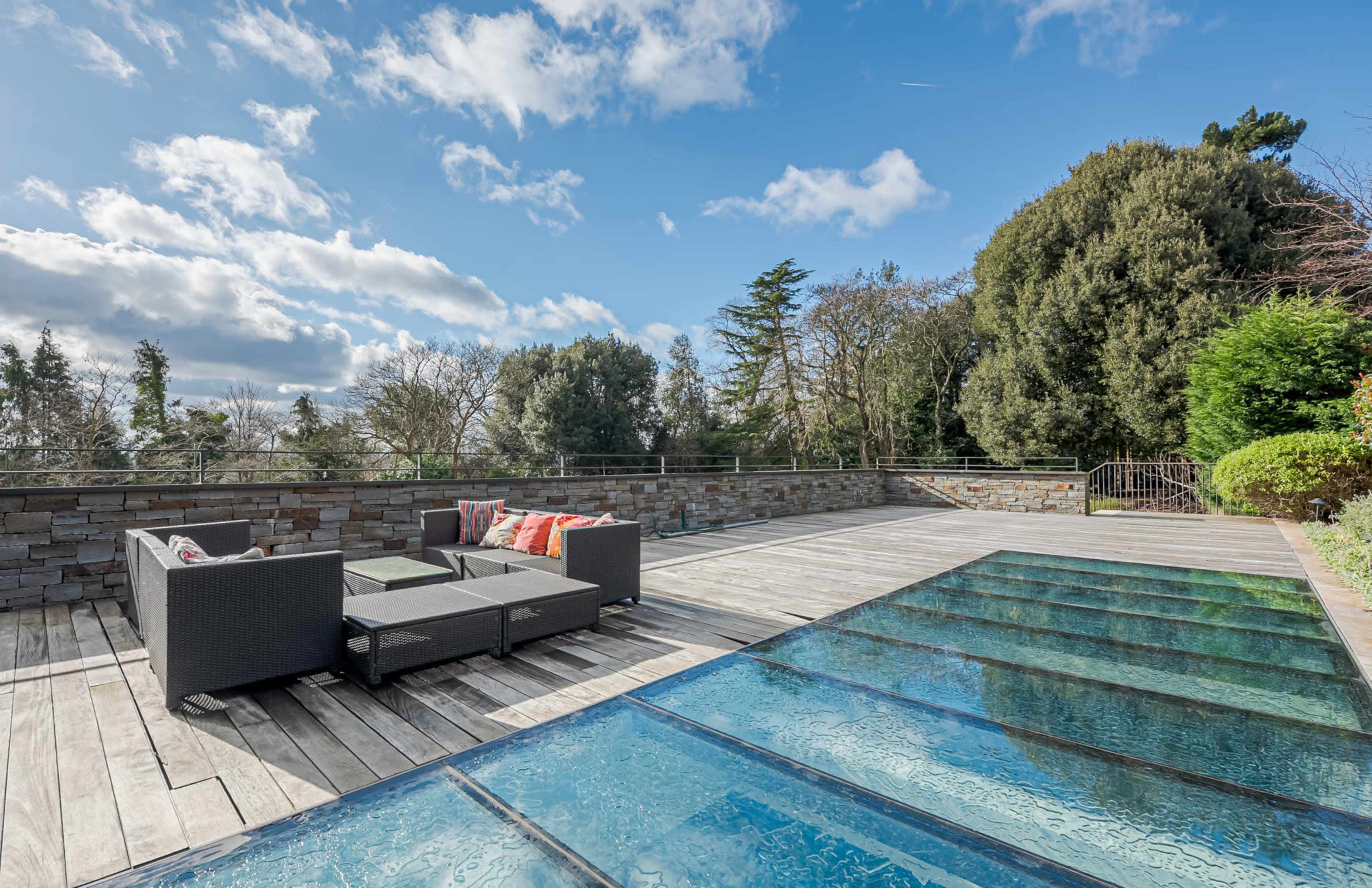 A modern patio features a glass-top area and a lounge set, surrounded by trees and greenery under a partly cloudy sky.
