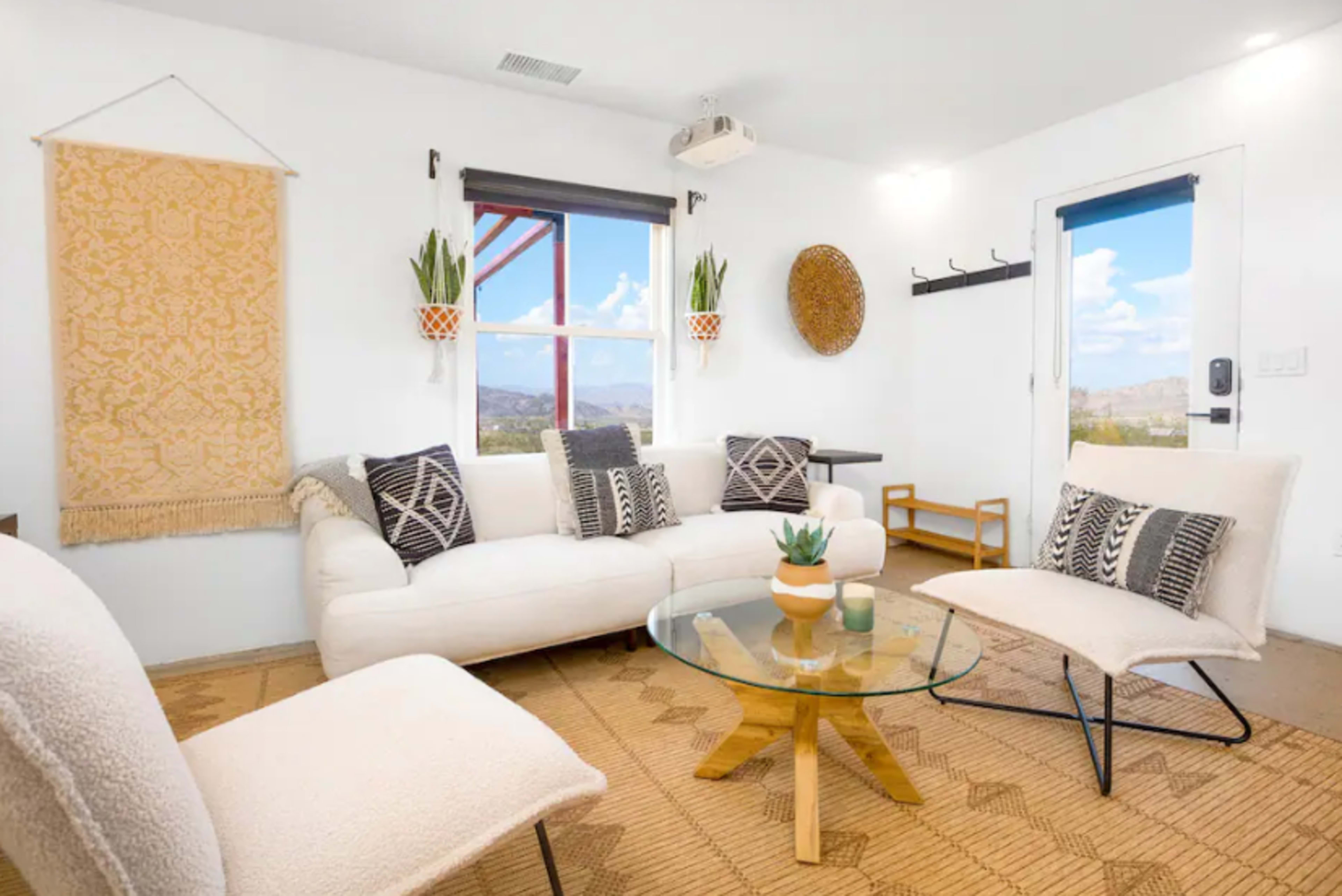 The image shows a bright living room with a white couch, glass coffee table, and two armchairs, set against a backdrop of large windows and decorative wall hangings.