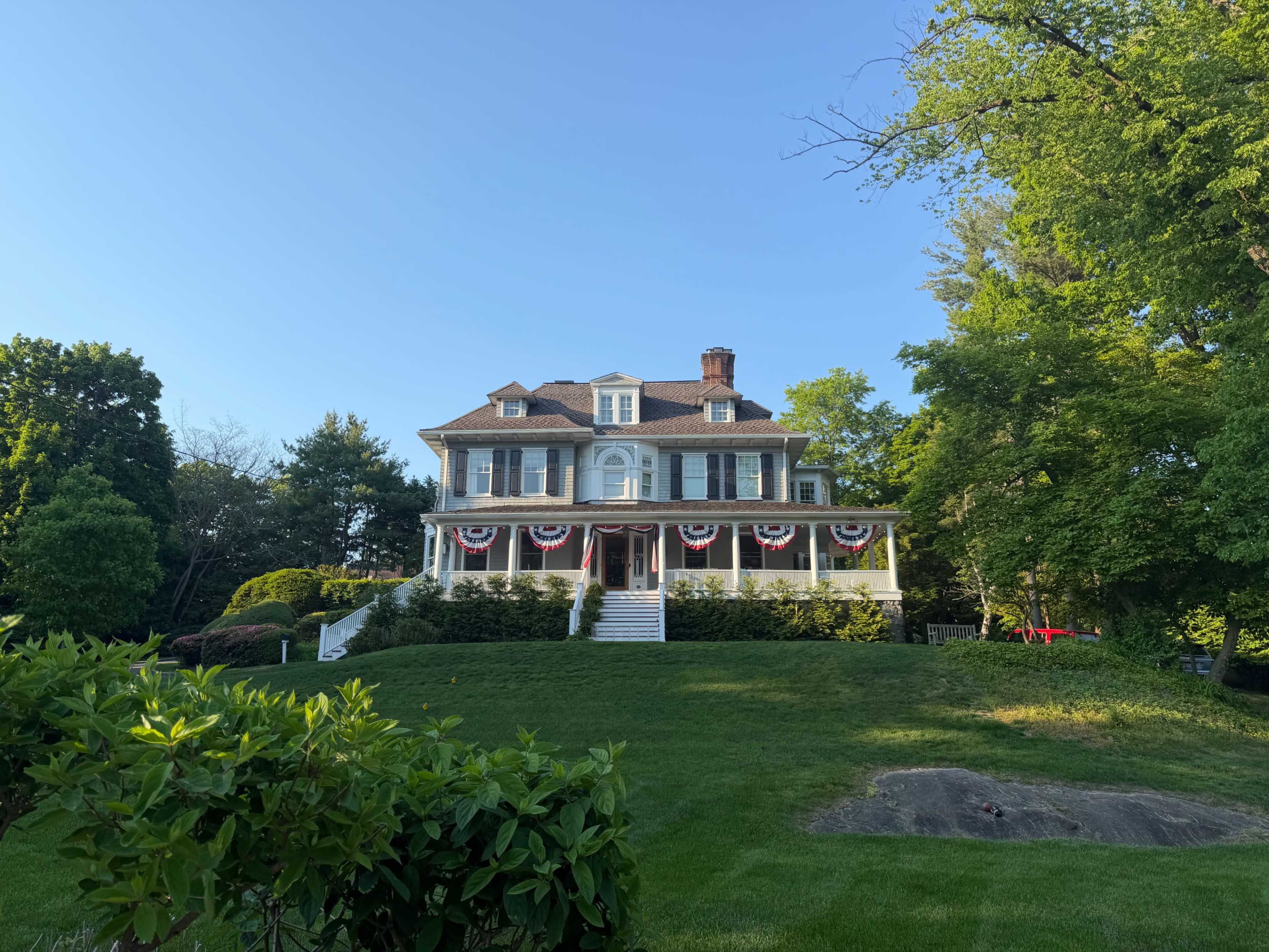A large, multistory house with a wraparound porch is surrounded by lush greenery and features decorative bunting along the railing.