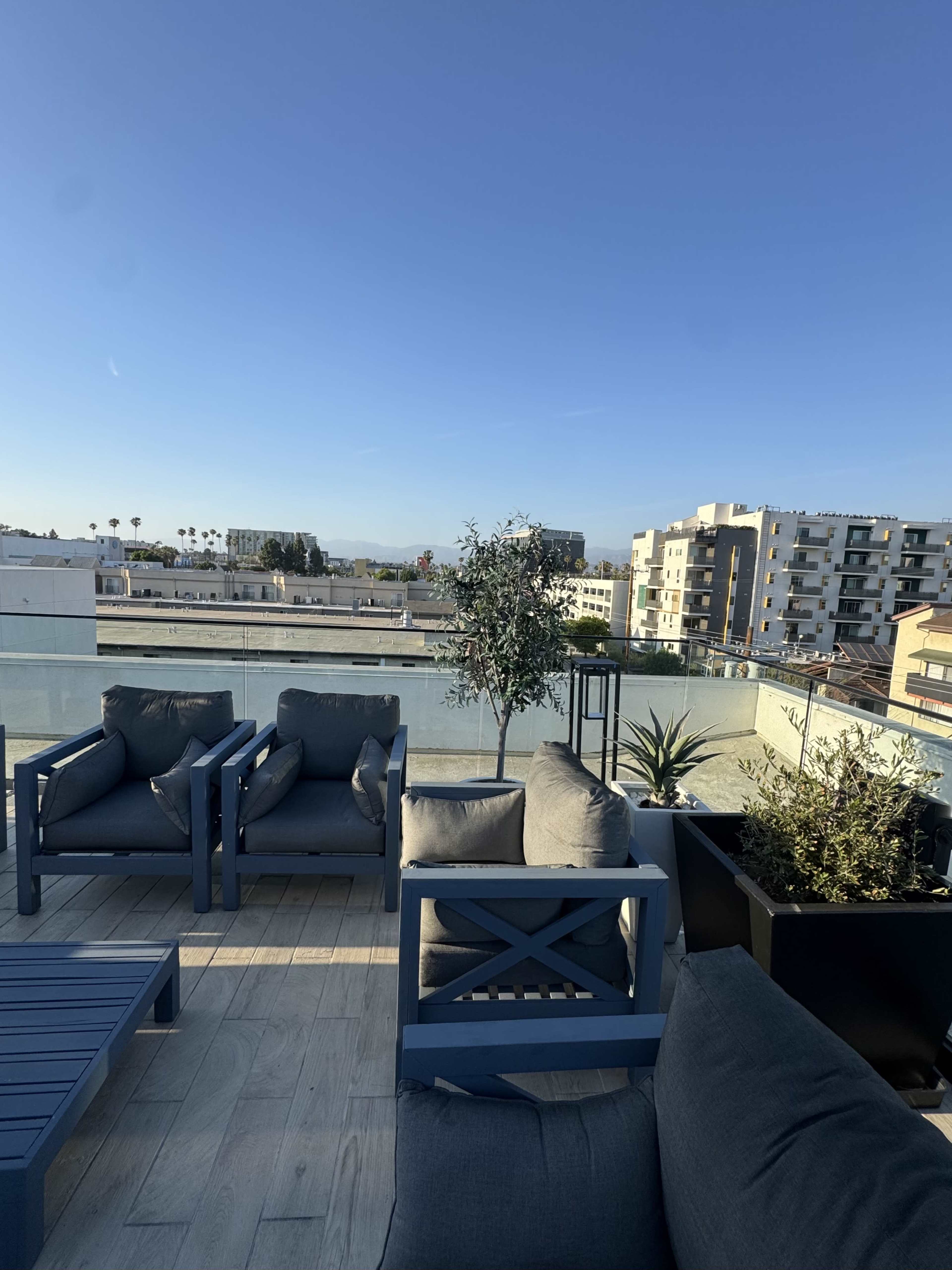 The image shows a rooftop seating area with blue outdoor furniture, overlooking a pool and urban buildings under a clear blue sky.