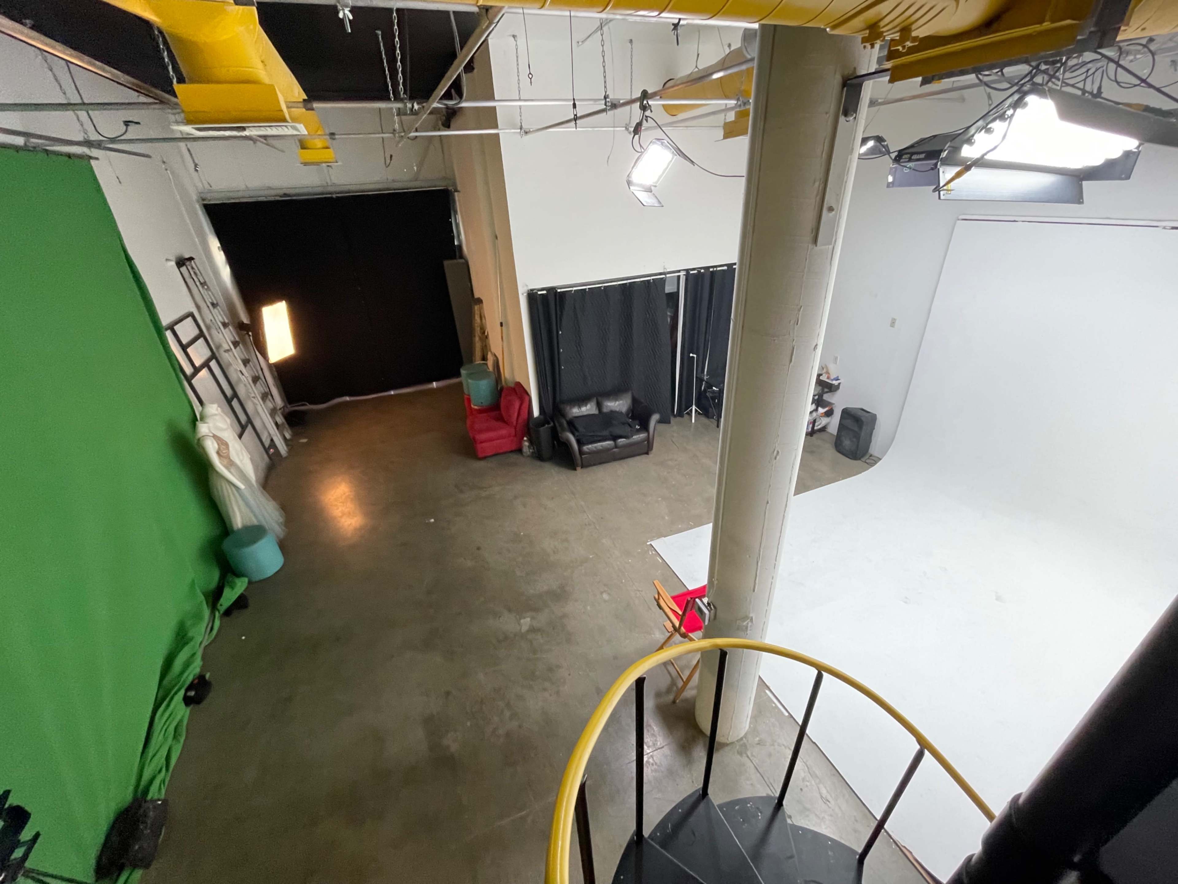 The image shows a spacious studio with a green screen, a white backdrop, and seating areas, viewed from a high angle near a spiral staircase.
