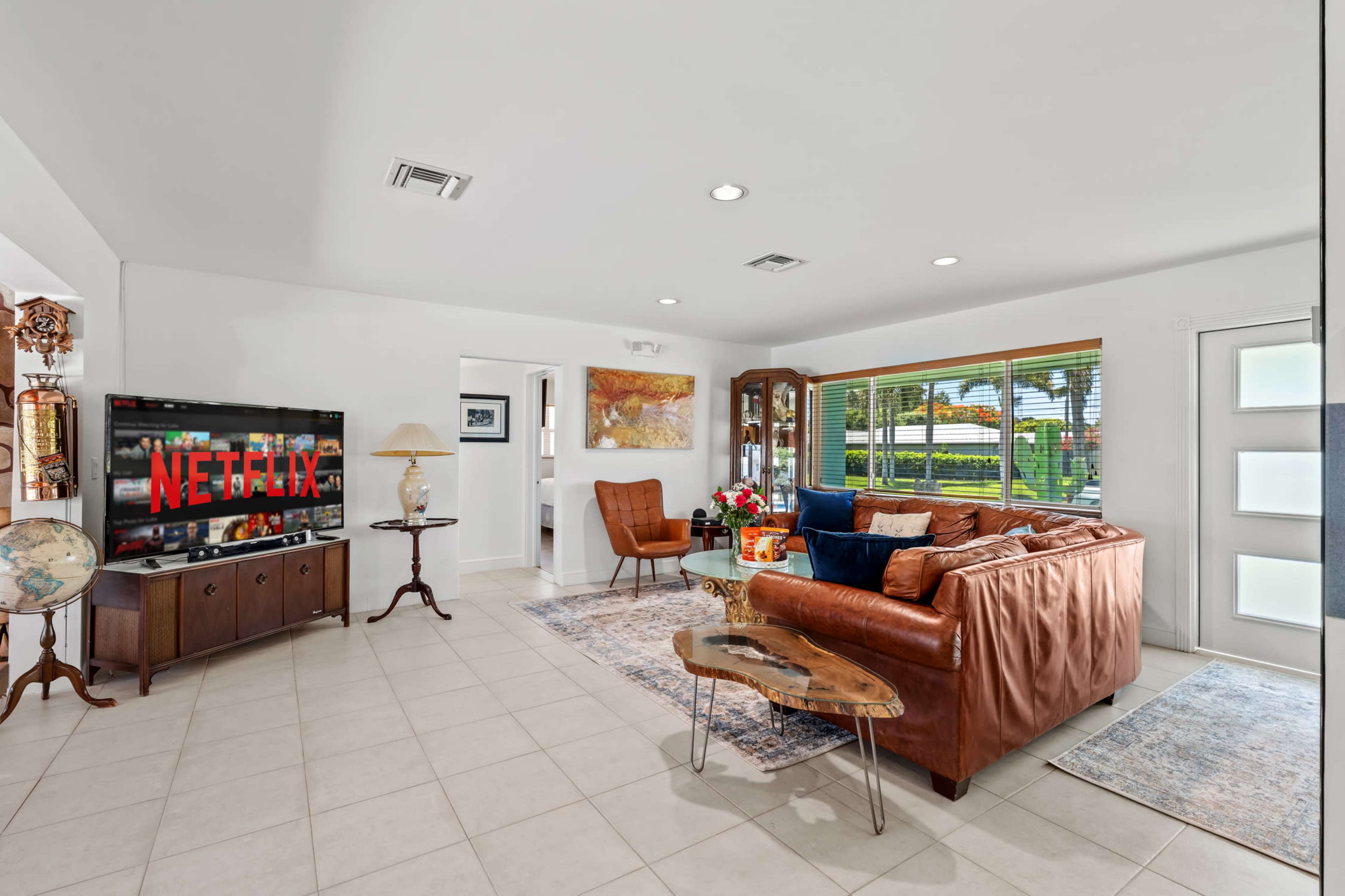 The image shows a modern living room featuring a brown leather sofa, a wooden coffee table, and a television displaying the Netflix logo, with large windows allowing natural light to enter.