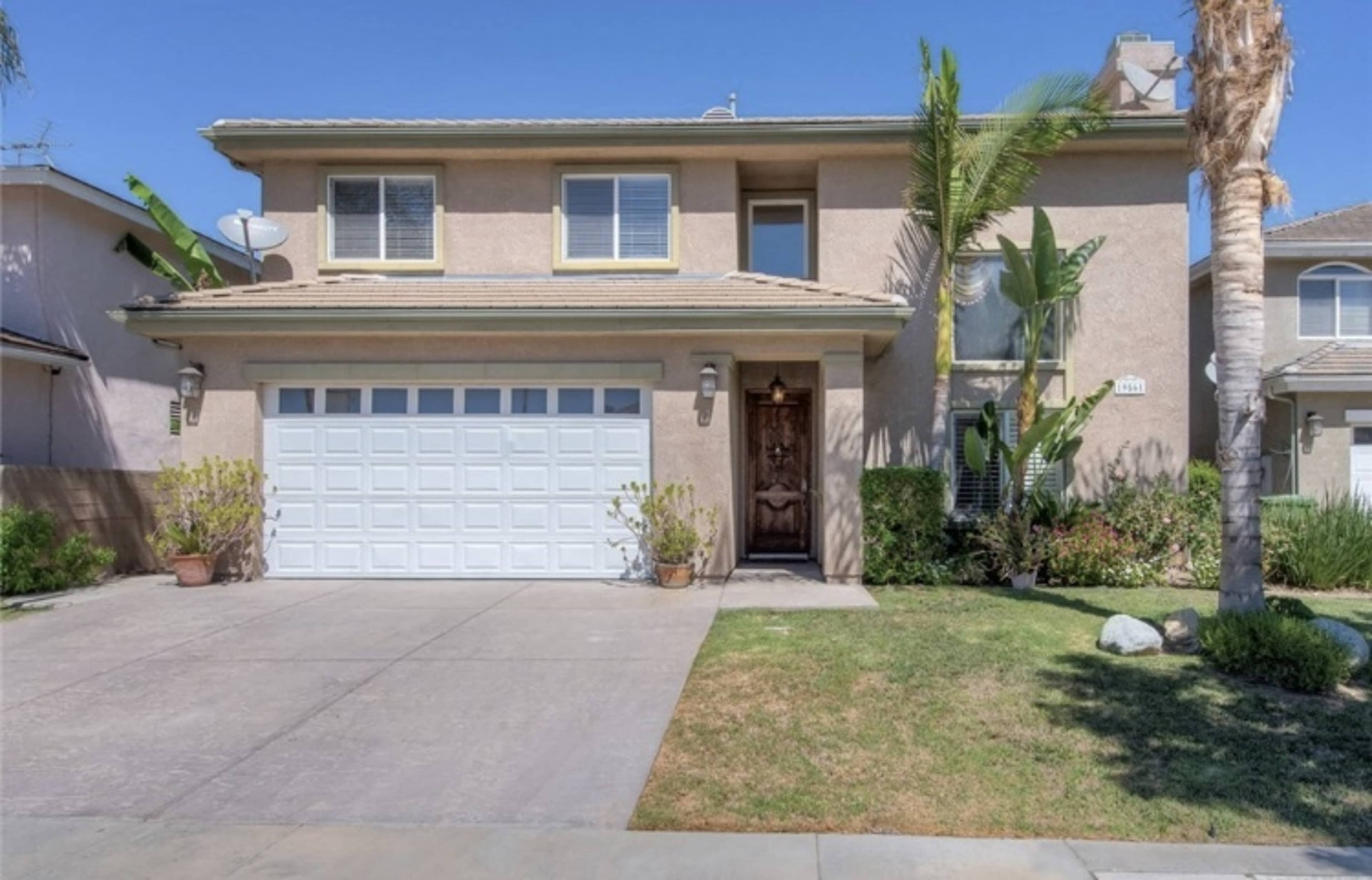 The image shows a two-story house with a garage and front yard, featuring a paved driveway and landscaping.