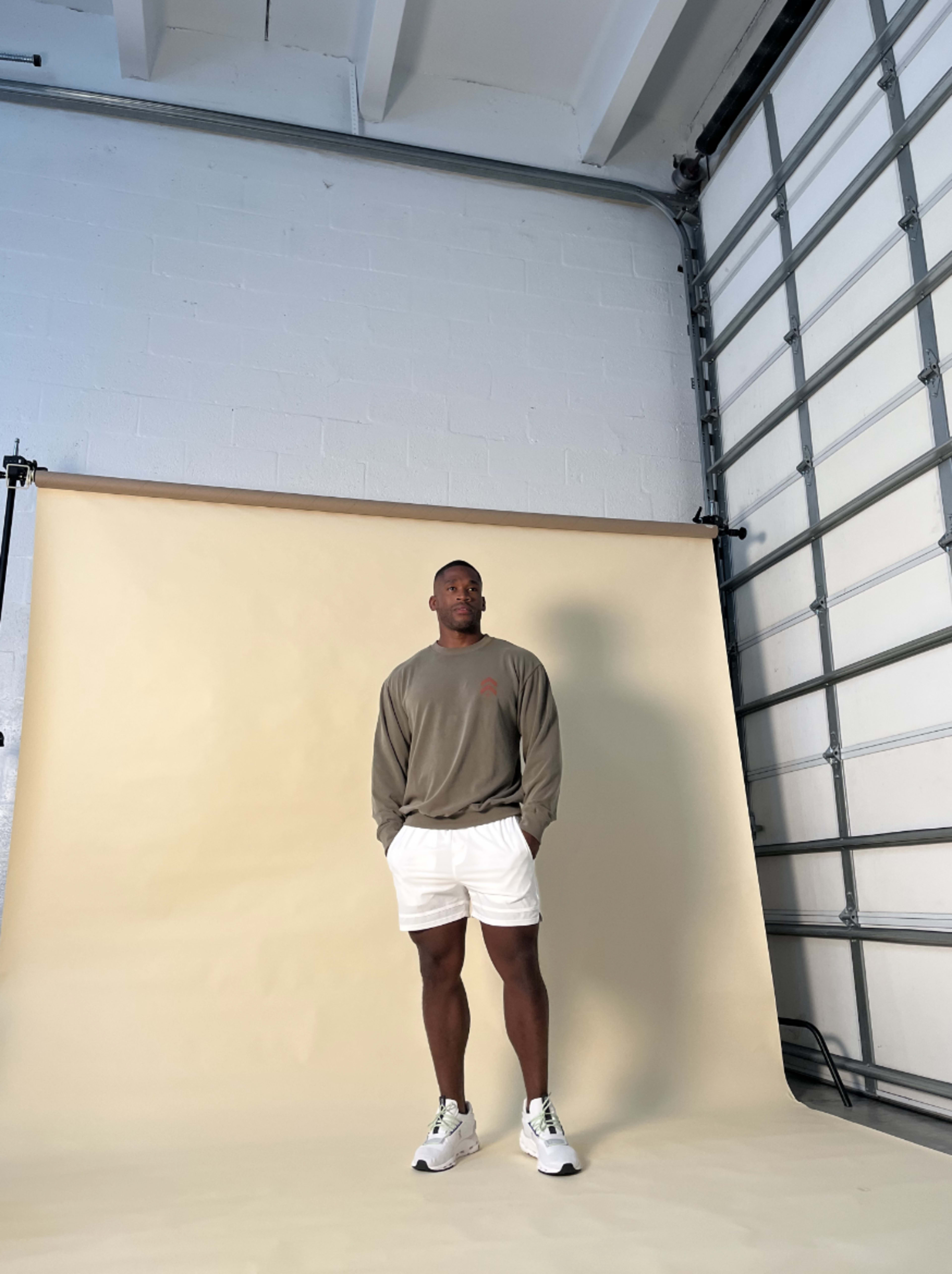 A person stands in a studio space with a beige backdrop, wearing a gray sweatshirt and white shorts.