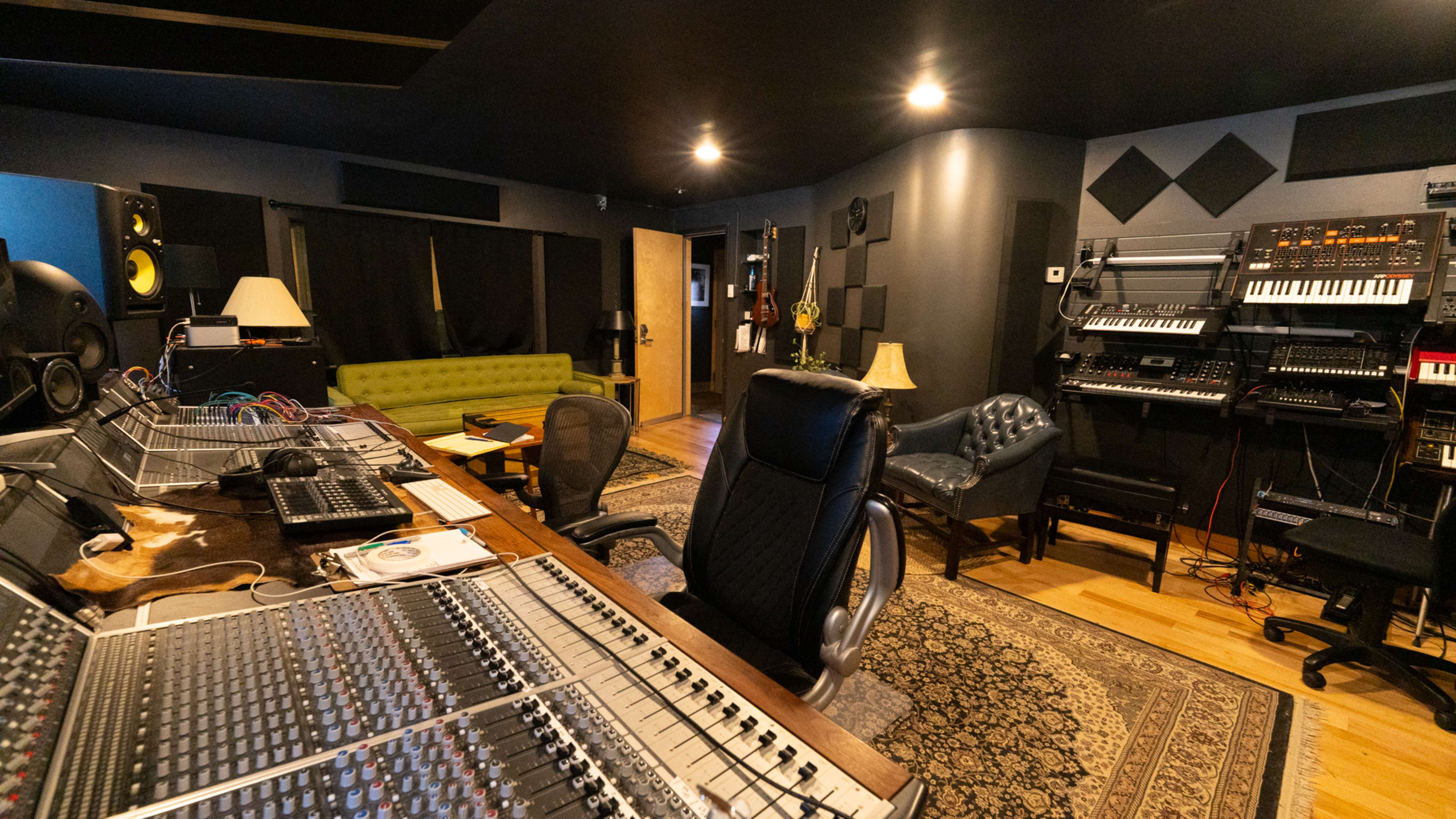 The image shows a music studio with a large mixing console, various electronic instruments along the walls, a green sofa, and a leather chair.