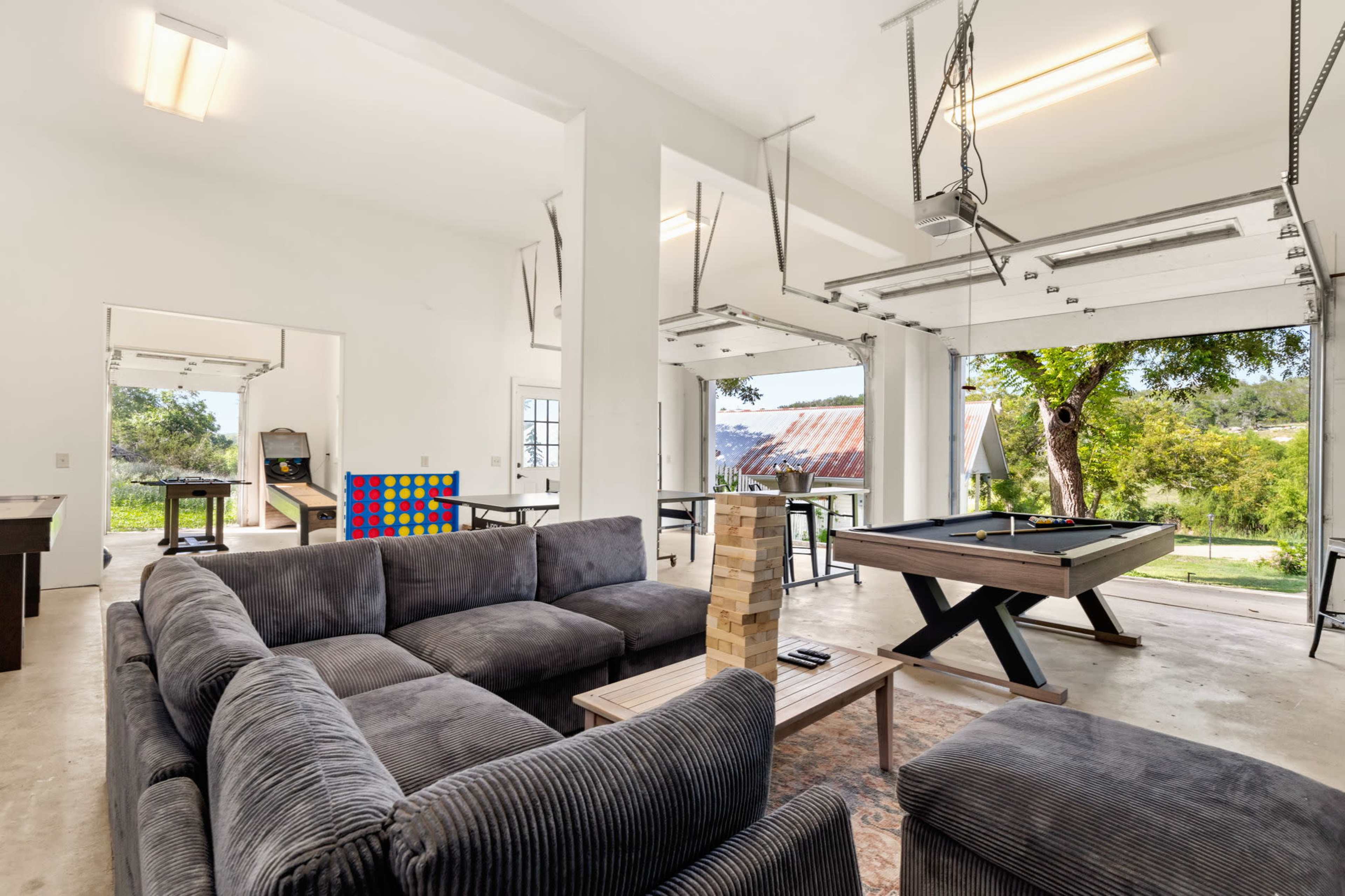 The image shows a spacious, open-concept room featuring a sectional sofa, a pool table, a colorful game board, and large garage doors that open to a view of the outdoors.
