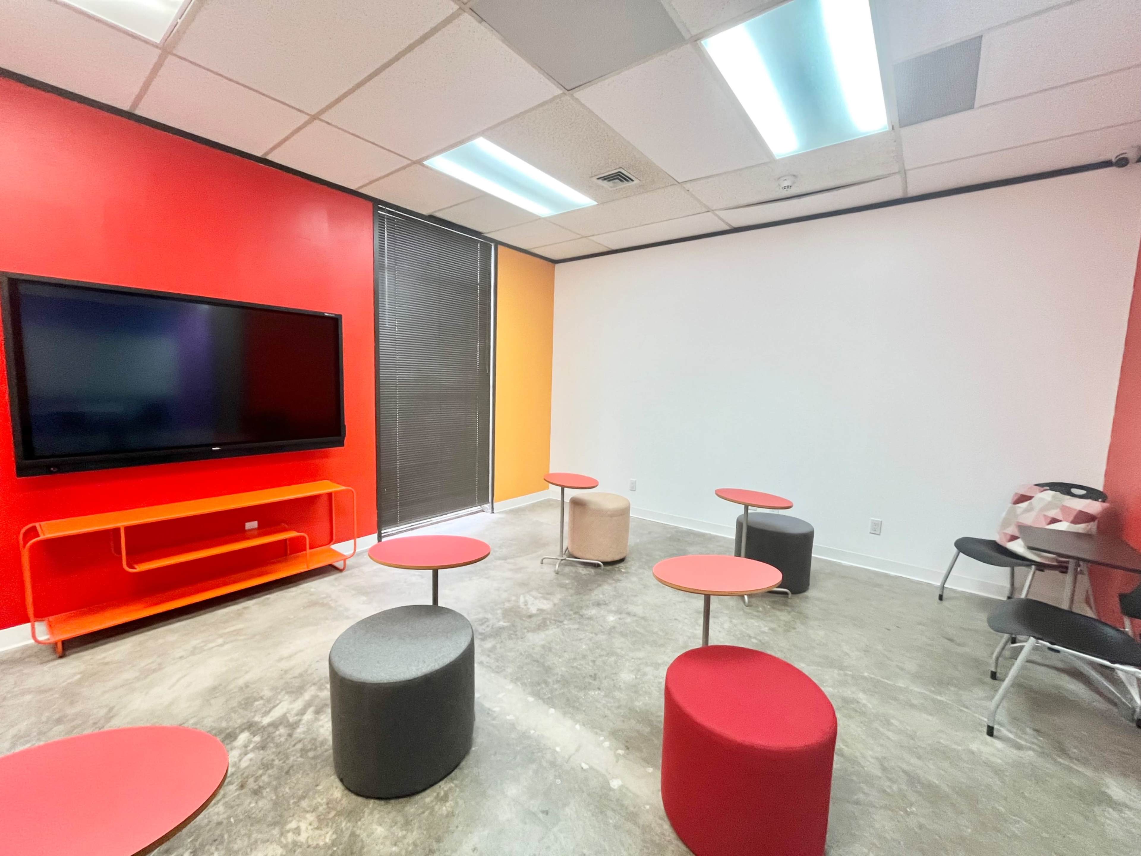The room features a television on a red wall, several round tables, and stools in various colors.