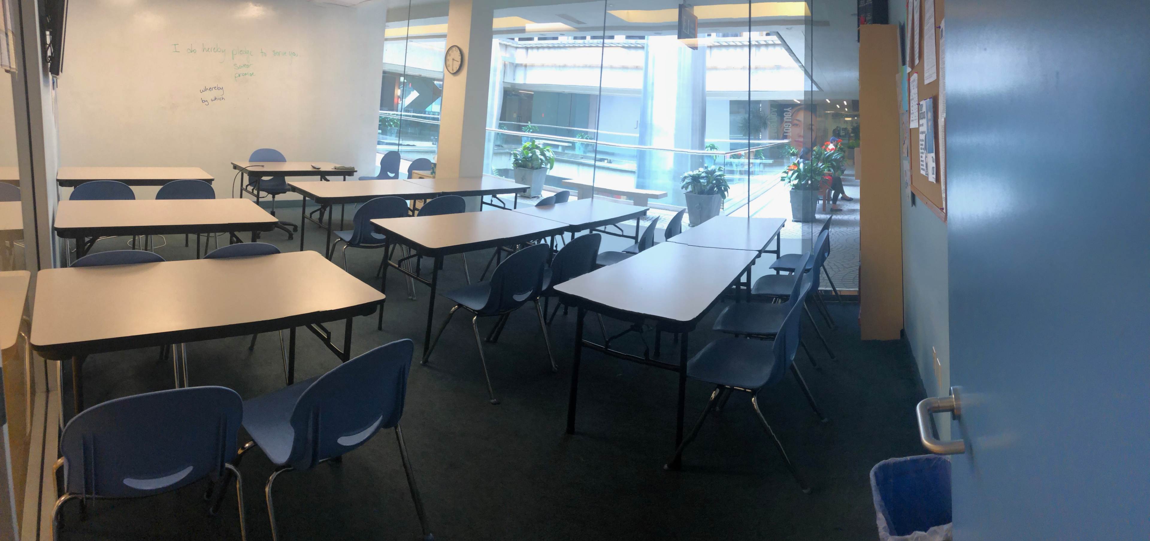 A classroom with multiple empty tables and chairs arranged in rows, overlooking plants and a courtyard through large windows.