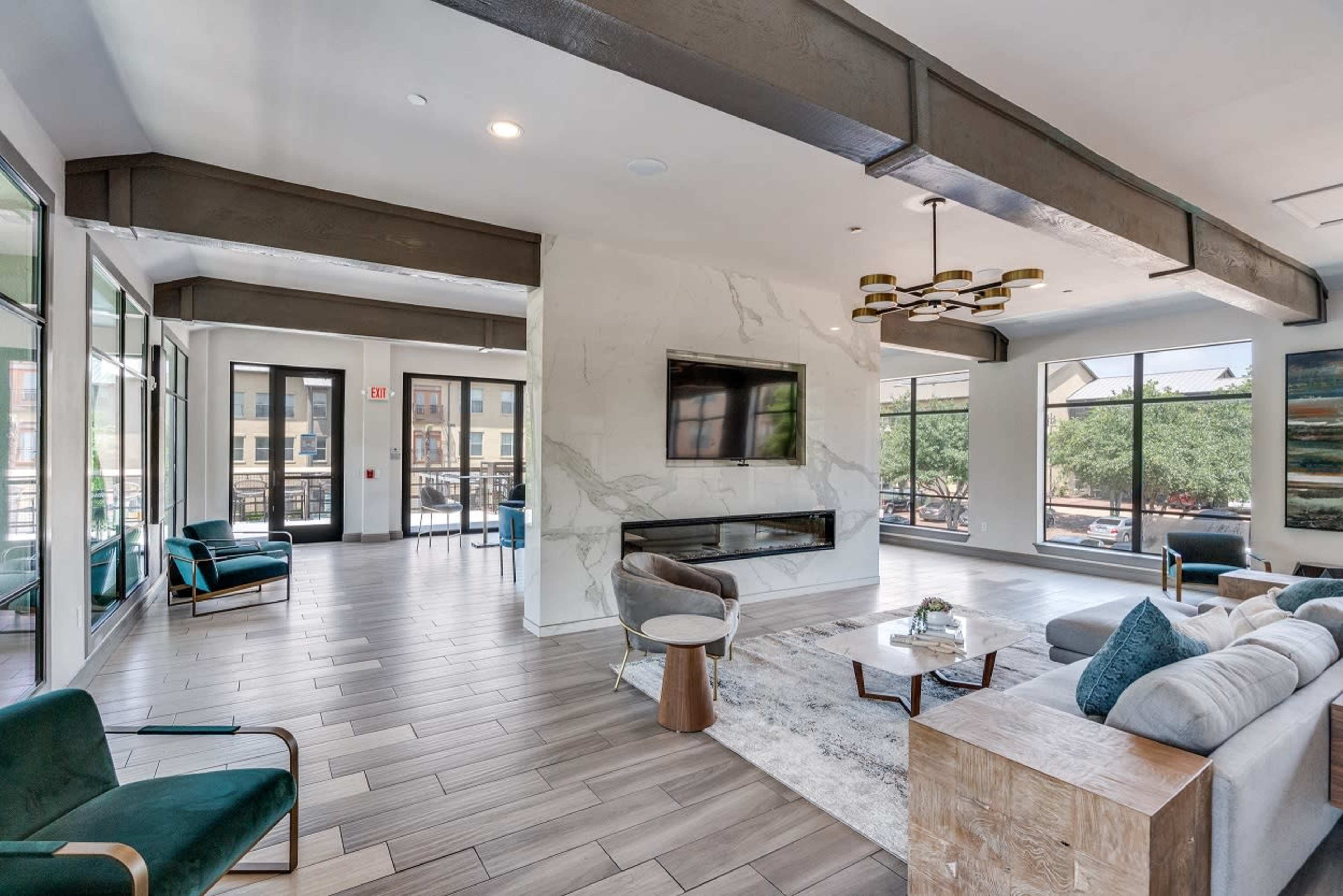 The image shows a modern living space featuring large windows, a central seating area with a sofa and chairs, a coffee table, and a wall-mounted television above a fireplace.