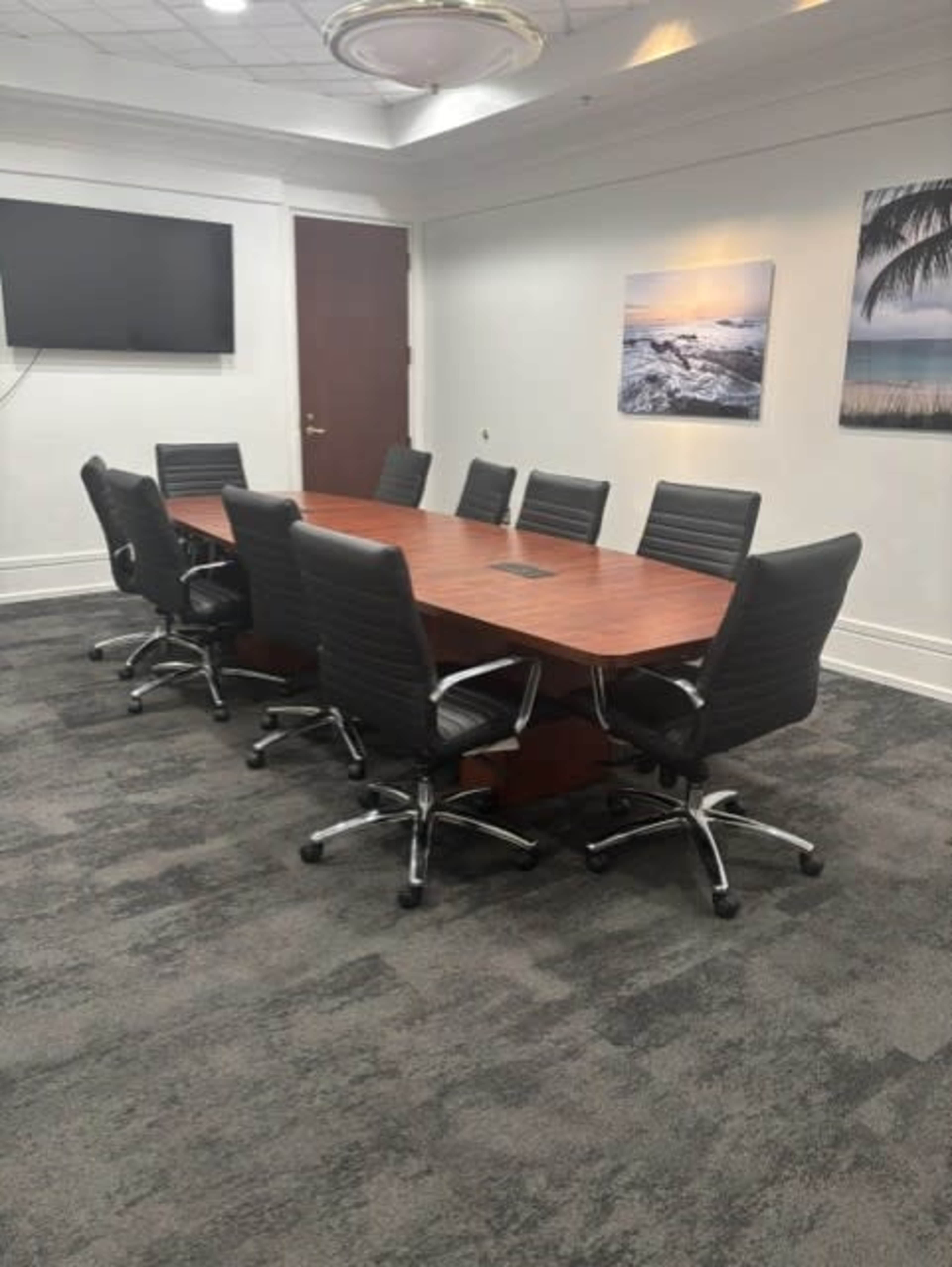 The image shows a conference room with a large wooden table surrounded by eight black swivel chairs and two framed pictures on the walls.
