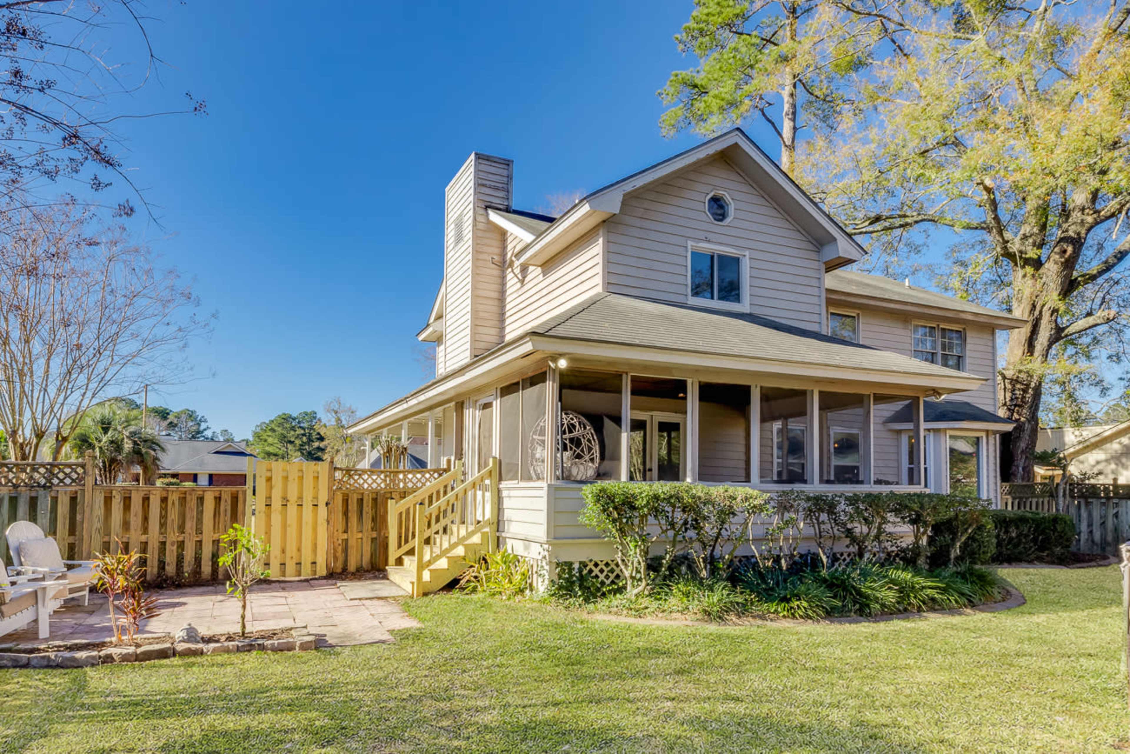 A two-story house with a screened porch and wooden stairs is surrounded by greenery and a fenced backyard.