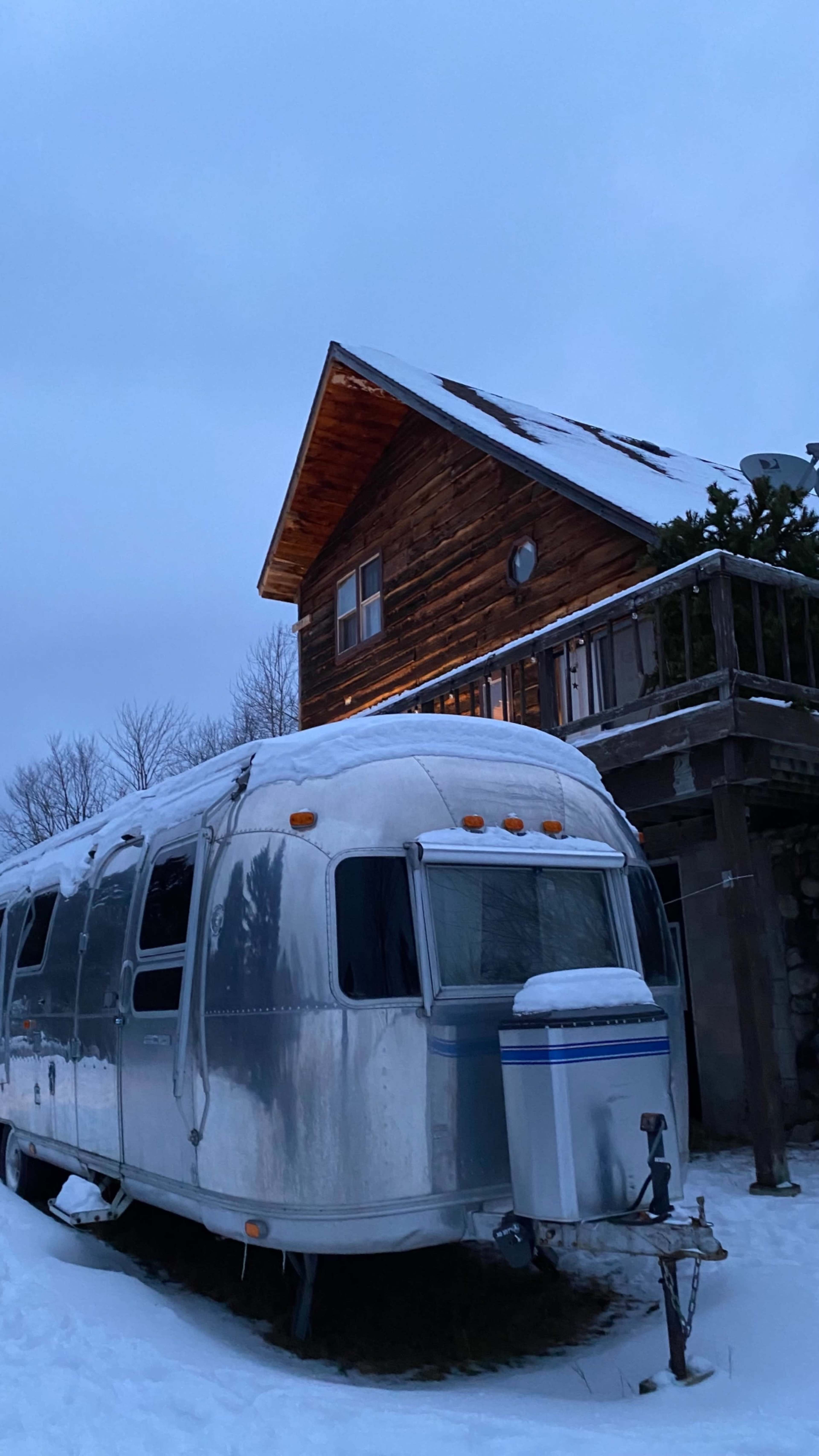 An Airstream trailer is parked in front of a wooden cabin covered in snow during twilight.