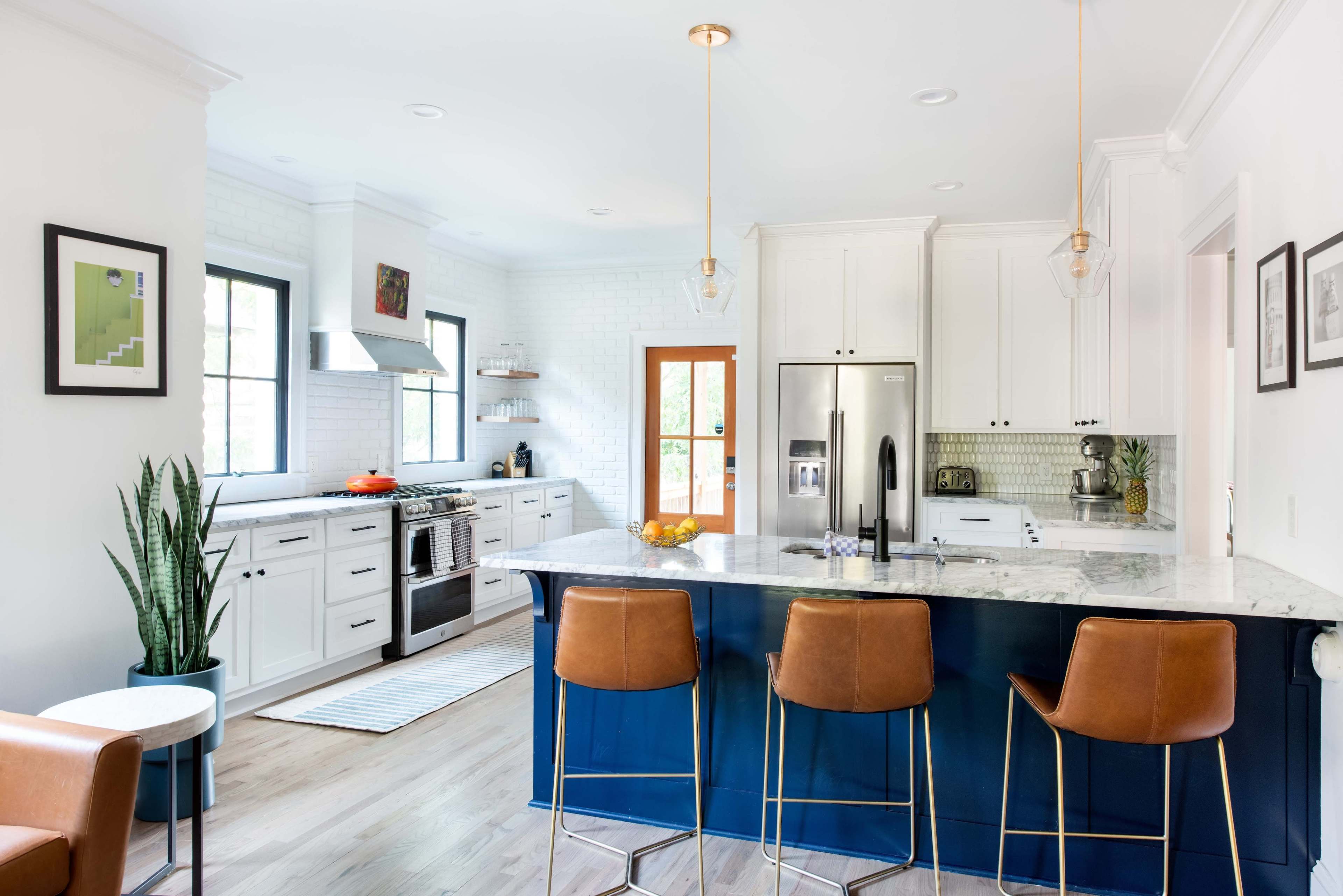 The image shows a modern kitchen with white cabinets, a marble countertop, stainless steel appliances, and four brown barstools at a blue island.