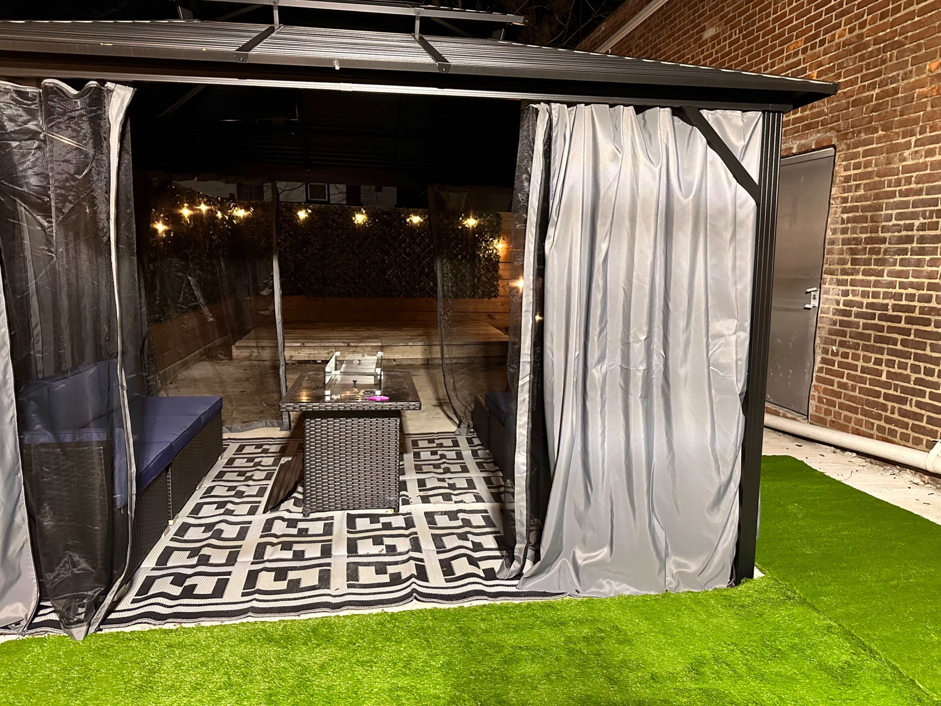 A shaded outdoor seating area with a black and white patterned rug and a glass table, enclosed by sheer curtains and surrounded by grass.